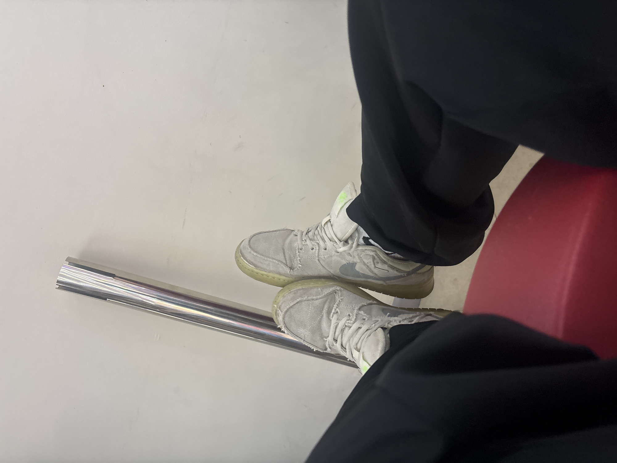 The image captures a first-person, downward view of a person's legs and feet, likely while seated. The subject is wearing loose-fitting black pants and a pair of light-colored, high-top sneakers.
The sneakers appear to be made of a distressed or frayed light grey/white fabric, giving them a worn look. They feature light green or yellow accents, possibly on the tongue or inner lining, and have translucent yellowish outsoles. A prominent Nike swoosh logo, in a slightly darker grey or silver, is visible on the side of the right shoe. The feet are elevated and resting on a shiny, cylindrical metal rail, which extends diagonally across the lower left portion of the frame. The rail appears to have some grooves or indentations near its end.
Part of a solid red object, likely a seat or a piece of furniture, is visible in the lower right and mid-right background, contrasting with the predominantly neutral tones. The background consists of a plain, light grey or off-white surface, which could be a wall or floor. The scene suggests an indoor setting where the person is resting.
There is no discernible text beyond the Nike swoosh. The location context provided is Yerevan, Armenia.
