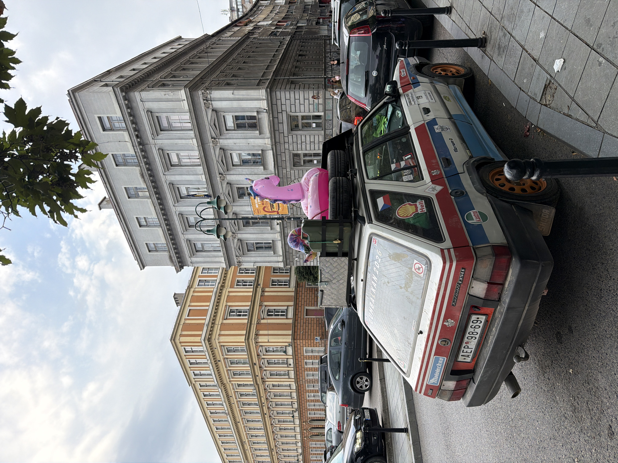The image captures a street scene in Sarajevo, Bosnia and Herzegovina, focusing on a modified white Skoda Favorit car with a pink inflatable unicorn on its roof rack. The car is parked on the right side of a street, with its rear facing the camera. It's adorned with various stickers and racing-style decals, including "GUMBALL 2023," "SHELL," "HELL," and "Kastrol." A Czech Republic flag sticker is visible on the driver's side rear window. The license plate reads "AEP 98-69."
Behind the car, several other vehicles are parked or in motion. In the background, prominent buildings with distinct architectural styles line the street. To the left, an orange and beige building with numerous windows and arched doorways stands out. To the right, a larger, grey building with ornate detailing and multiple stories dominates the skyline. The sky above is partially cloudy. The street is paved with asphalt, and a sidewalk with stone paving is visible to the right of the car. Several black bollards line the edge of the sidewalk. The overall atmosphere suggests an urban environment.