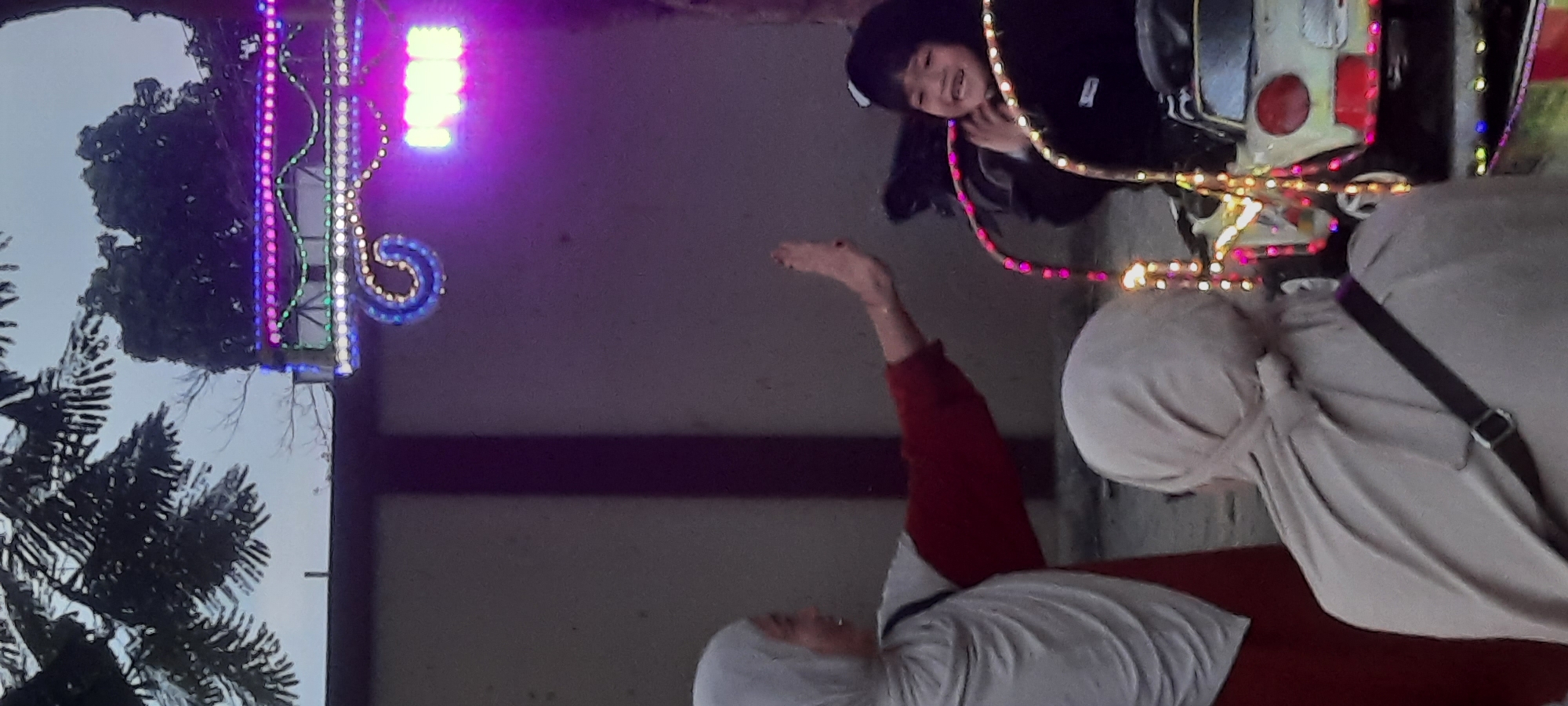 In Kanigoro, Indonesia, a vertical, medium shot captures two individuals with a child engaged in an activity. On the left, a person wearing a light-colored hijab and a red top extends their right hand, palm facing outwards, in what appears to be a high-five gesture. To the right of this individual, a child, wearing a dark jacket and a black hat, is seated in a small, car-shaped ride adorned with colorful lights. The child is looking up and smiling, with their hands near their face. In the foreground and partially obscured, another person in a light-colored hijab is visible from the back, facing away from the camera. The background features a muted, greyish sky with the silhouette of trees and branches, along with a structure decorated with neon-colored lights. The overall scene suggests a recreational setting, possibly a fair or amusement park, during twilight or overcast conditions.