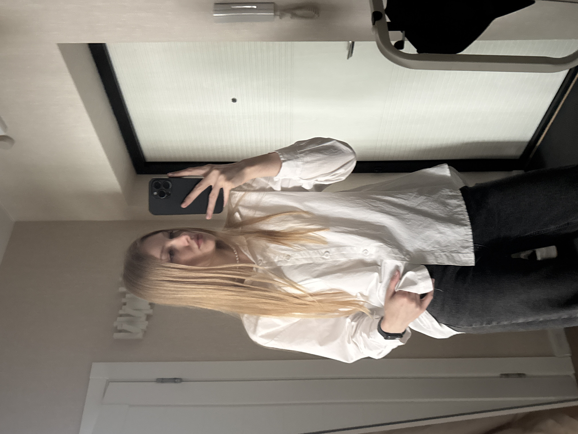 A blonde woman is taking a selfie in what appears to be a bathroom or dressing area. She is wearing a white button-down shirt and dark jeans. She is holding a smartphone in her left hand to take the picture. The reflection in the mirror behind her shows part of the room, including a door and what looks like a clothing rack. Based on visual evidence, there is no information in the image to determine that this image was taken in Perm, Russia.