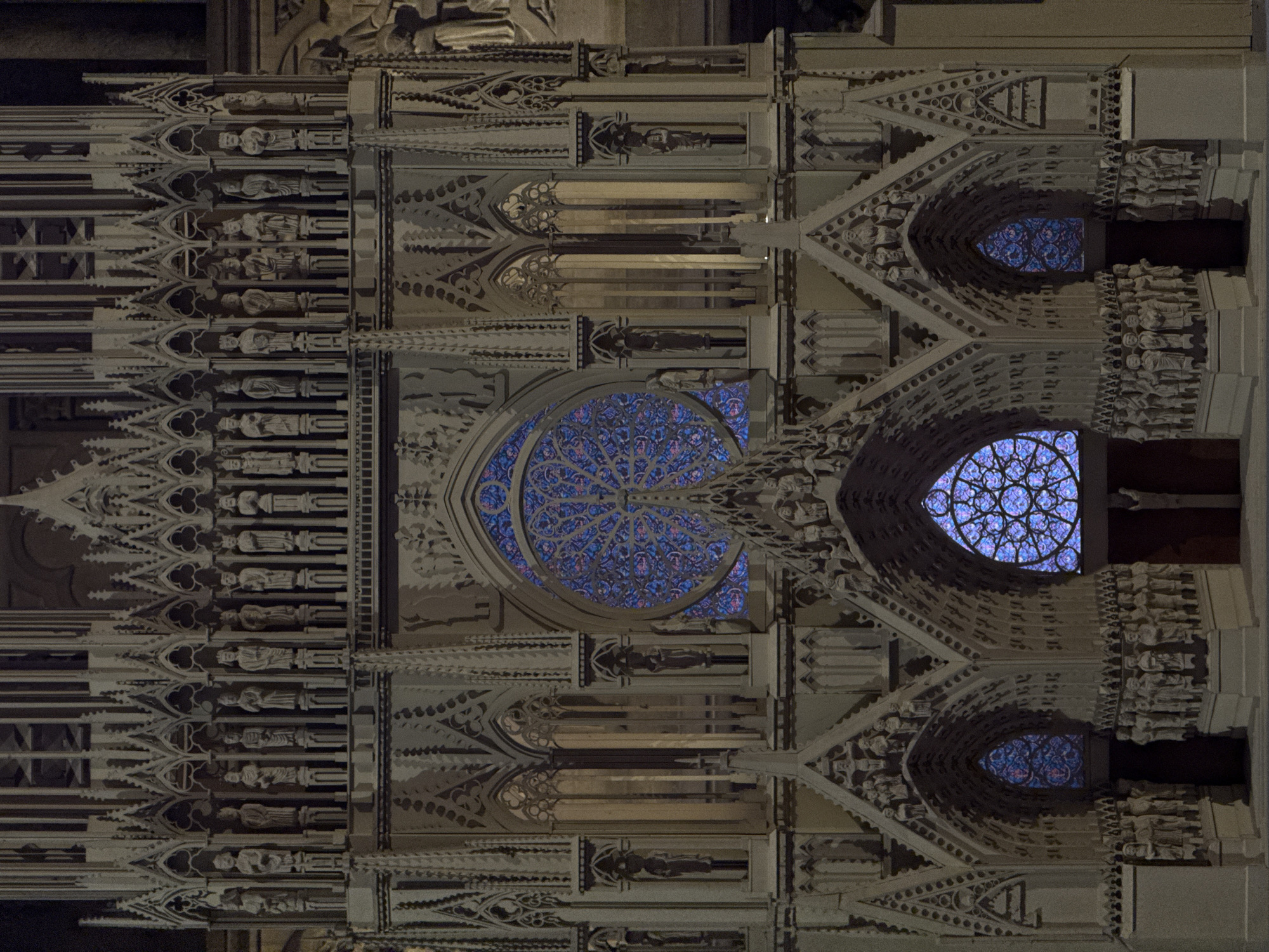 This media file presents a close-up view of the intricately detailed Gothic facade of a cathedral at night, most likely Reims Cathedral in Reims, France, given the context. The image is rotated 90 degrees clockwise from its natural orientation.
The scene is dominated by the elaborate architectural and sculptural elements of the building, illuminated by artificial lights against a dark night sky. Three prominent stained-glass rose windows glow vibrantly with blue, purple, and hints of red. The largest of these is centrally located, showcasing complex geometric patterns in its tracery. A smaller rose window appears to its right, and an even smaller one further to the right and slightly below, all emitting light from within.
Numerous sculpted figures, possibly saints, kings, or other religious personages, adorn the facade. They are arranged in vertical tiers along the left side of the composition, standing in niches and beneath canopies. More sculptures are visible framing the rose windows and integrated into the arches and architectural details throughout.
The cathedral's stone, likely limestone, appears in shades of grey and muted beige under the artificial illumination, creating strong contrasts with the dark background and the luminous stained glass. Elaborate pointed arches, pinnacles, and delicate stone tracery are visible, showcasing the high level of craftsmanship typical of Gothic architecture. No specific activity or event is depicted, emphasizing the architectural grandeur of the structure itself. There is no visible text.