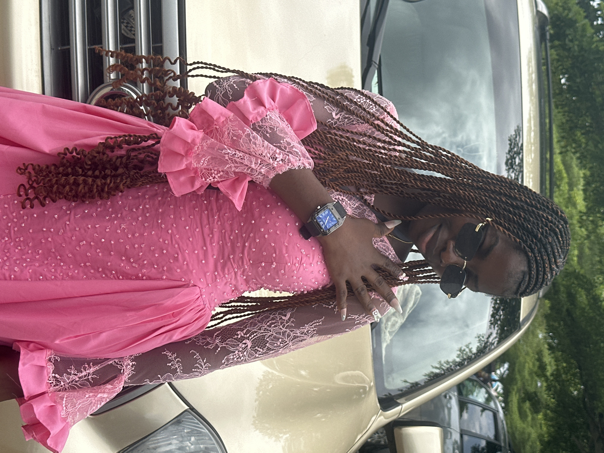 In Itoga, Nigeria, a young Black woman stands in front of a gold SUV. She wears a long-sleeved pink dress adorned with small, scattered beads and lace accents on the sleeves. Her hair is styled in numerous brown braids, some of which hang down her front. She has on black sunglasses and a silver watch on her left wrist. Her right hand is raised, with her fingers resting on her chest and a ring visible on her index finger. The background shows greenery and other vehicles, suggesting an outdoor location.