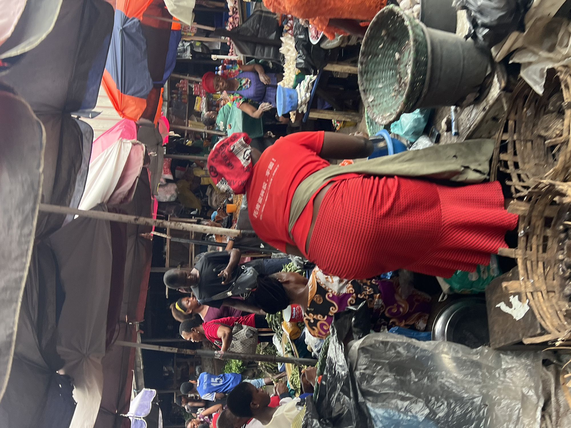 The image depicts a bustling outdoor market scene in Nkpor, Nigeria. The market is covered by a variety of colorful awnings and umbrellas, suggesting an active marketplace. In the foreground, a woman in a bright red shirt and red skirt is visible from the back, with a green apron draped over her shoulder. She appears to be standing behind a stall or display. To her left, a person in a patterned dress is seated.
The market is filled with people, some of whom are actively engaged in transactions or preparing goods. A young man in a blue jersey with the number 15 is seen in the background, facing away from the camera. Other individuals are interspersed throughout the scene, some conversing, others tending to stalls.
Various items are on display and for sale, including what appear to be fresh produce like green vegetables and cucumbers. Baskets of varying sizes, some made of woven material, are present, likely used for carrying or displaying goods. There are also plastic containers and bags, as well as what looks like raw meat or fish on a table in the mid-ground. The overall atmosphere is one of lively commerce, characteristic of a busy Nigerian market.