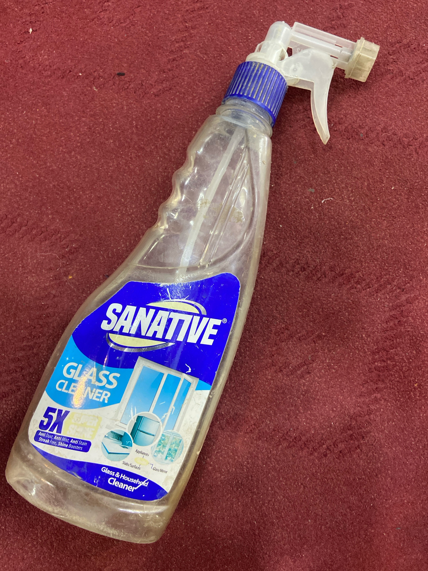 A clear plastic bottle of "SANATIVE GLASS CLEANER" with a blue spray nozzle and blue and white label lies on a textured, dark red surface. The label features a graphic of a window and the text "GLASS CLEANER 5X". The bottle is partially filled with a clear liquid. The red surface appears to be carpet or fabric, with visible fibers and some dust or lint. The scene is lit from above and to the left, casting slight shadows. The image was taken in the city of Lahan, Nepal.