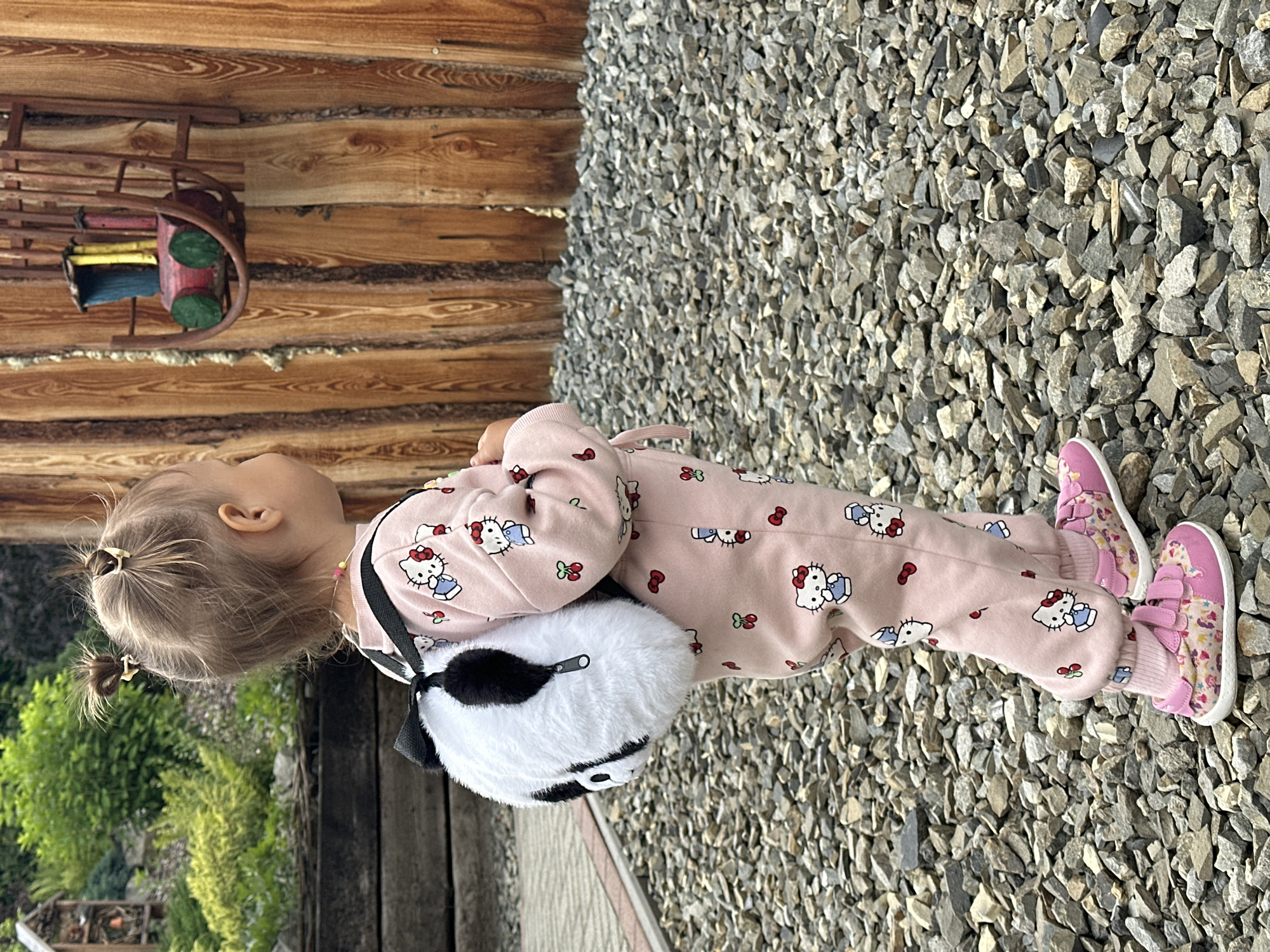 A young child, viewed from the side and from the knees down, is standing on a surface of gray gravel. The child is wearing a pink pajama set adorned with Hello Kitty characters and cherries. On their feet are pink sneakers with colorful patterns and velcro straps. The child is also carrying a fluffy, round, black and white bag that resembles a panda face. Behind the child, to the left, is a rustic wooden wall made of logs. On the wall hangs a small, decorative wooden sled with red, yellow, green, and blue accents. The scene is outdoors, likely in a rural or natural setting, given the wooden structures and gravel. The lighting suggests it is daytime, possibly late afternoon given the warm sunlight casting shadows. The child's posture and the presence of the toy suggest a moment of leisure or play. The visible text is "Hello Kitty" on the pajamas. The location context provided is Wisła, Poland.