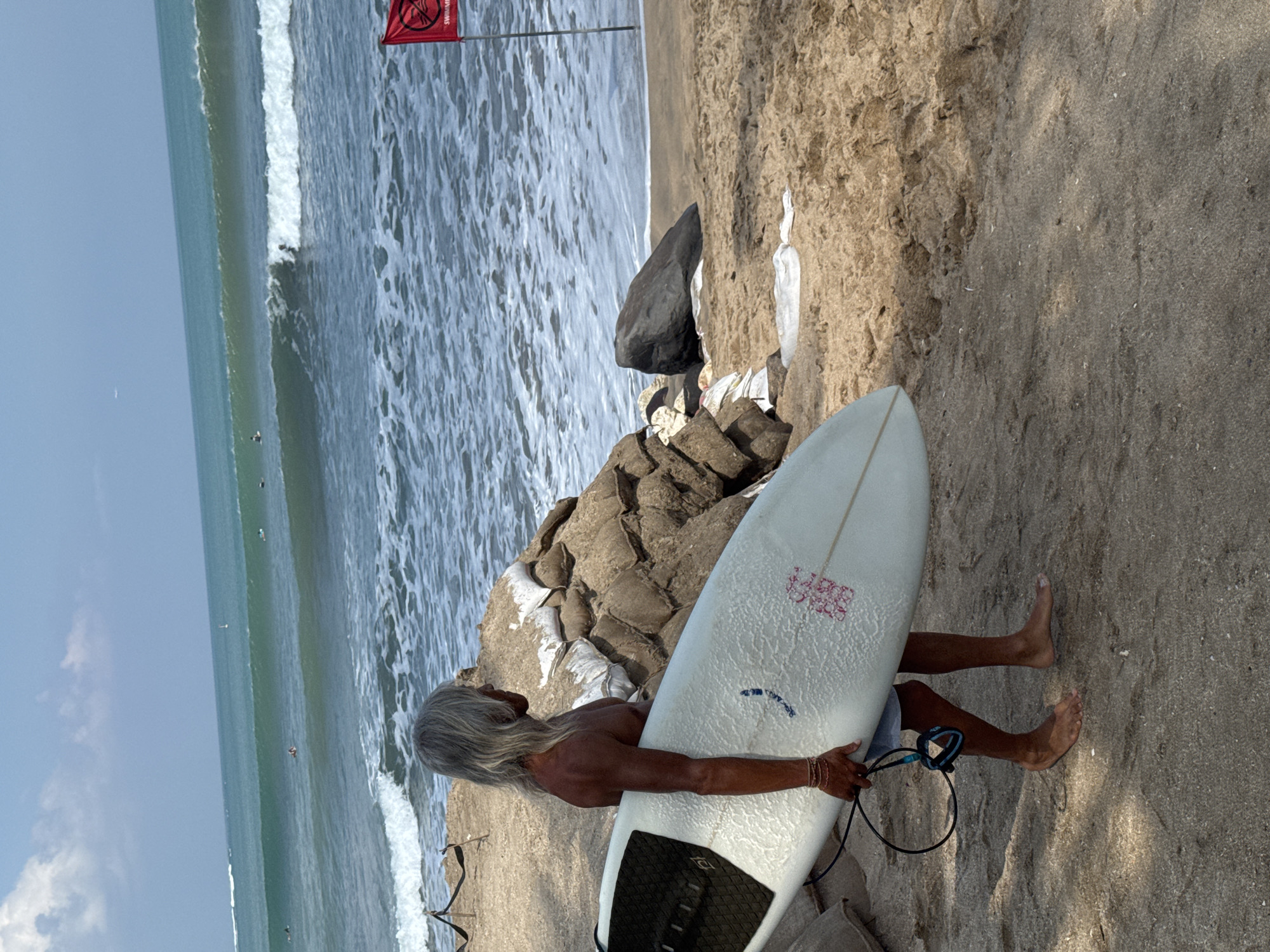 A shirtless man with long gray hair stands on the beach in Kuta, Indonesia, carrying a white surfboard. He is facing right, looking out at the ocean, where waves are breaking and several surfers are visible. Sandbags are piled behind him, and a red flag with a prohibition symbol is planted in the sand to his right. The sky is blue with scattered clouds, and the ocean is a deep turquoise. The sand is light brown and textured.