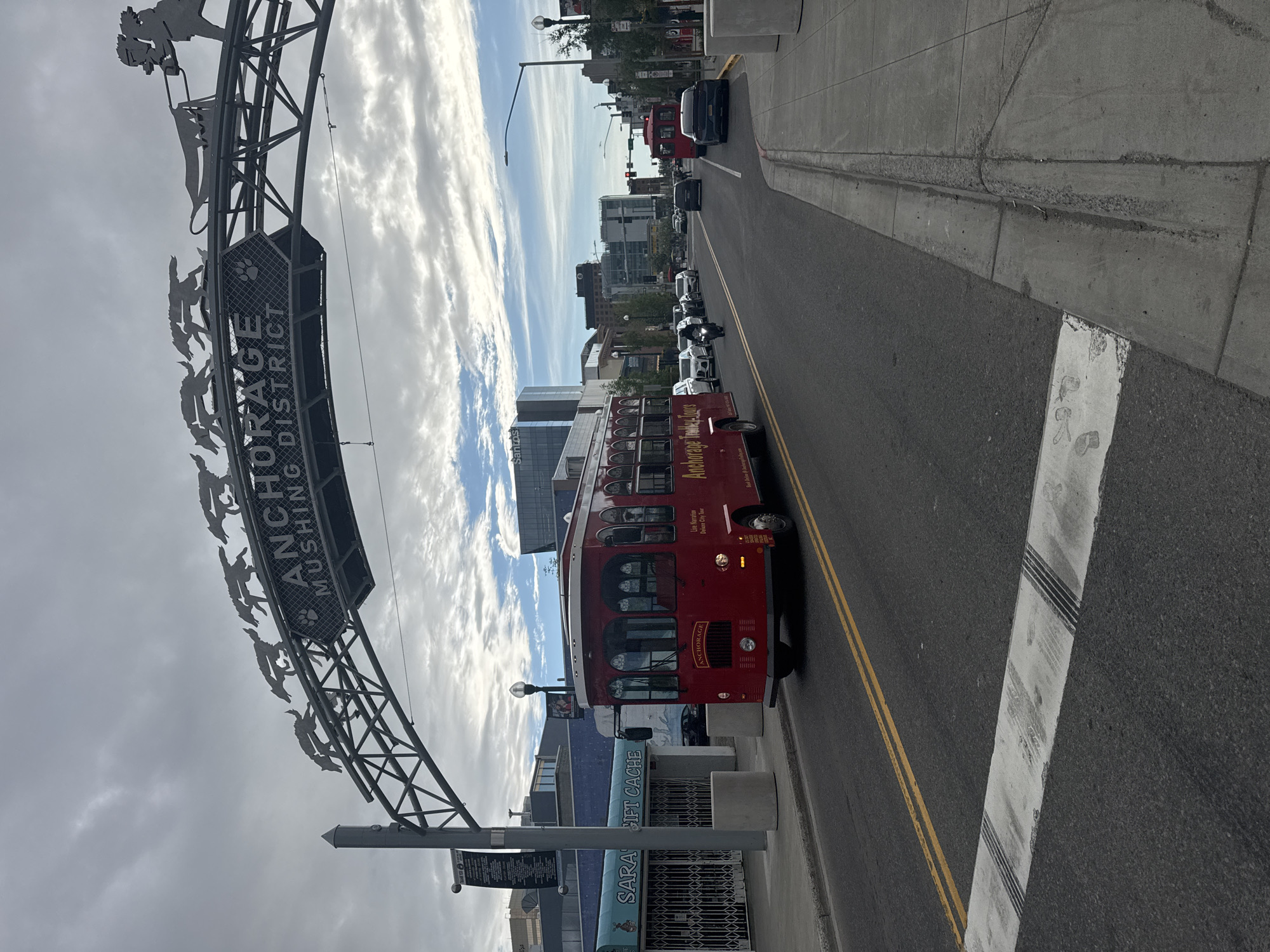 The image shows a street scene in Anchorage, Alaska. A red trolley bus with "Anchorage Trolley Tours" written on its side is stopped at the side of the road. The trolley has multiple windows and is adorned with ornate metalwork. A large metal archway with the words "ANCHORAGE MUSHING DISTRICT" is visible to the left of the trolley. The archway features a silhouette of sled dogs pulling a sled. The street is paved and has yellow double lines dividing the lanes. Other vehicles, including cars and a motorcycle, are seen on the road. Buildings line the street, and the sky is partially cloudy, suggesting it is daytime. The overall scene depicts a tourist-oriented area in an urban setting. Visible text also includes "Santos" on a building and "SARAH GIFT CACHE" on a storefront.