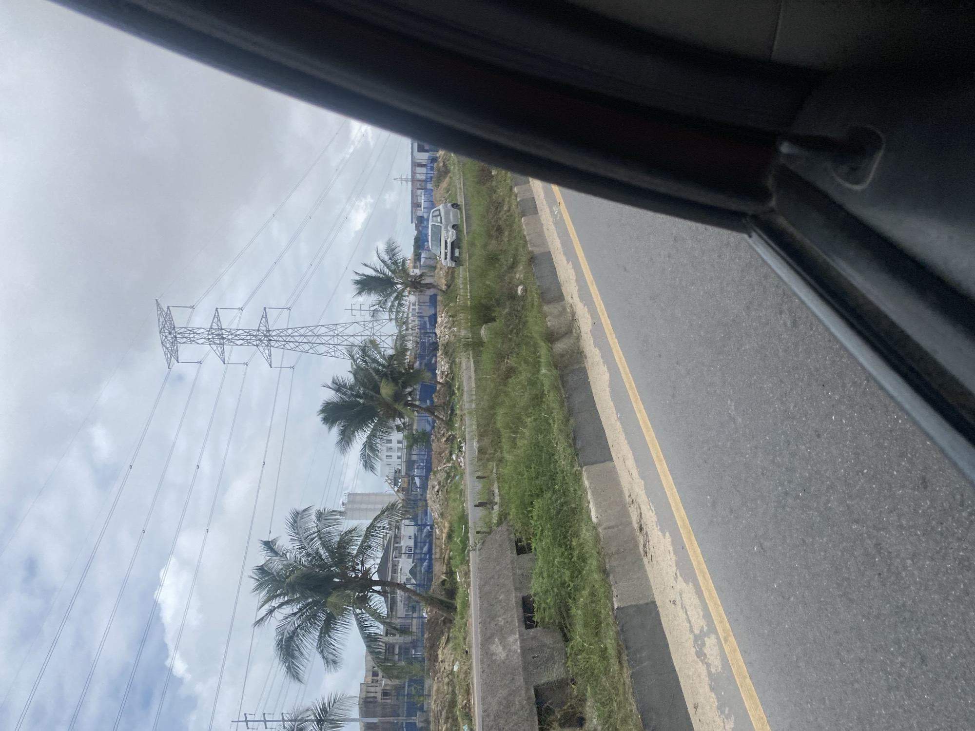 The image captures a roadside scene, likely in Ilado, Nigeria, characterized by a stretch of asphalt road with a yellow dividing line. On the left side of the road, a concrete barrier separates the pavement from a grassy embankment. Beyond the embankment, several palm trees are visible, with some appearing to lean. In the background, there are construction sites with unfinished buildings and industrial structures, including a prominent electricity pylon. A small, light-colored car is parked on the right side of the road, amidst the vegetation. The sky is overcast with clouds. The perspective suggests the image was taken from inside a vehicle, with a portion of the car's interior visible in the foreground on the right side of the frame.