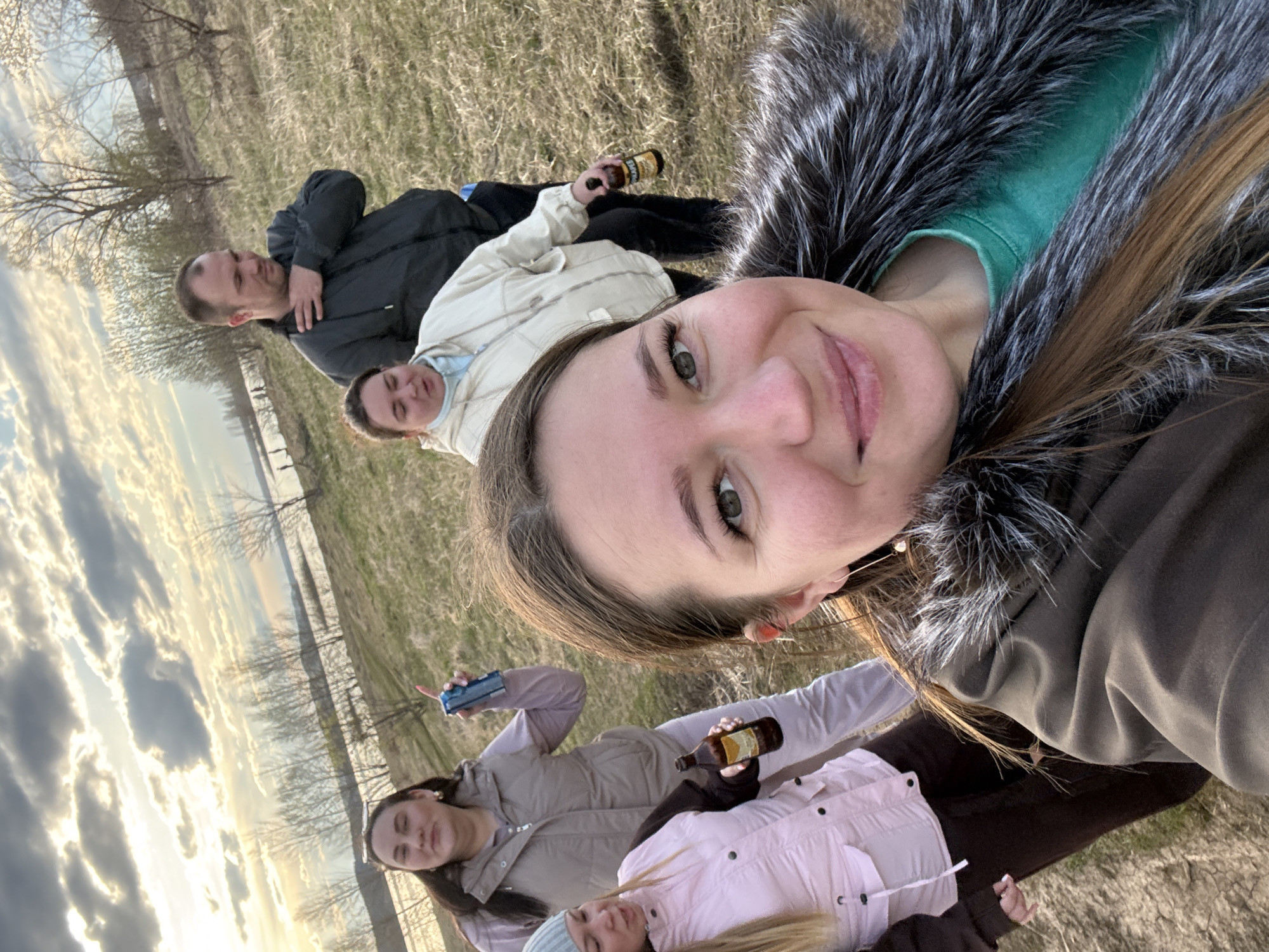 A woman in a fur-trimmed parka takes a selfie with five other people in a field. The field is near a body of water and trees. Some people are holding cans and bottles. The photo was taken in Шолья, Russia.