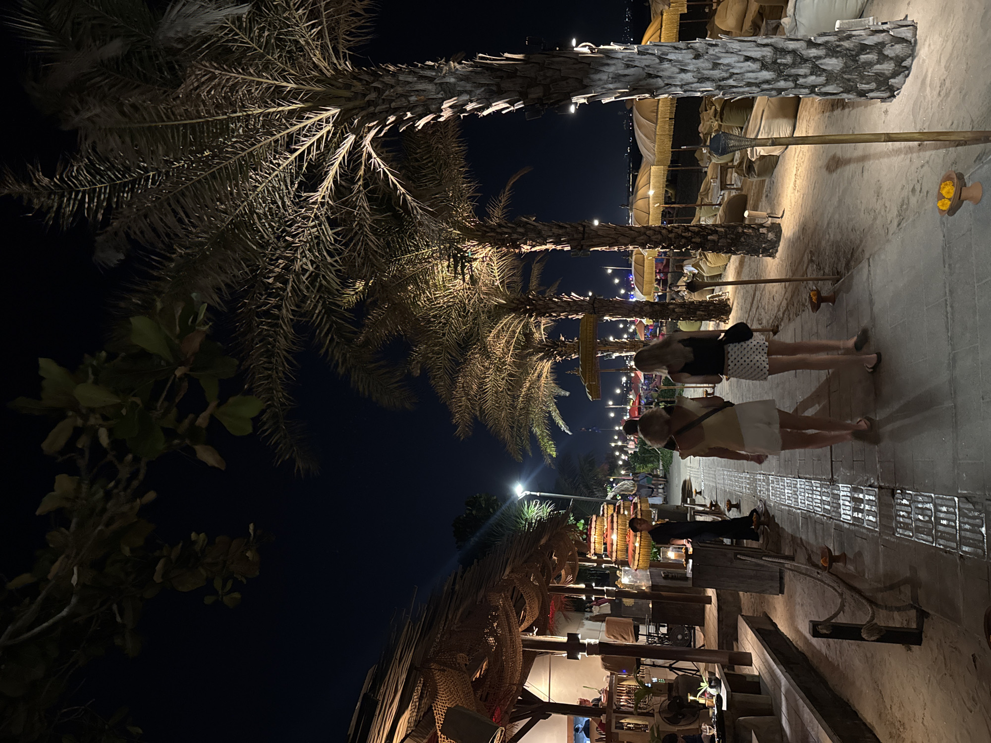 In the city of Kuta, Indonesia, at night, two women walk on a paved path beside a sandy beach lined with palm trees. The woman in the foreground wears a light yellow top and white shorts, while the woman behind her wears a black top and a white skirt with black polka dots. They are walking away from the viewer towards a beachside establishment with a thatched roof, where lights and decorative nets are visible. The scene is illuminated by artificial lights, casting shadows on the ground and highlighting the textures of the palm trees. Beach chairs and umbrellas are set up on the sand. A man in a black uniform stands near a tiered display of what appears to be fruit or flowers. The sky is dark, indicating nighttime.