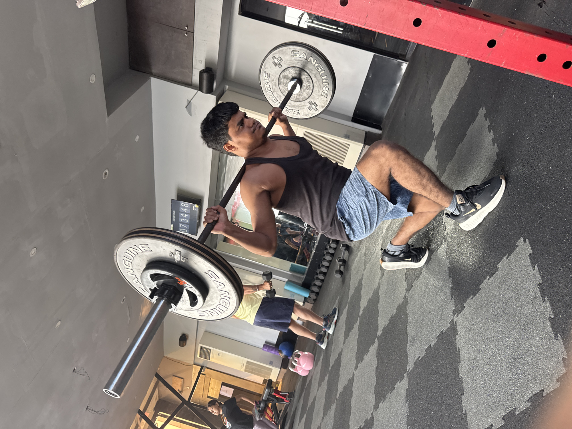 The image shows a man in a tank top and shorts performing a barbell exercise in a gym. The barbell is loaded with weight plates labeled "Sanguine". Another person can be seen in the background using dumbbells. Various other gym equipment is visible, including weight racks and kettlebells. The floor has a diamond-patterned rubber mat. The location is identified as Kolkata, India.