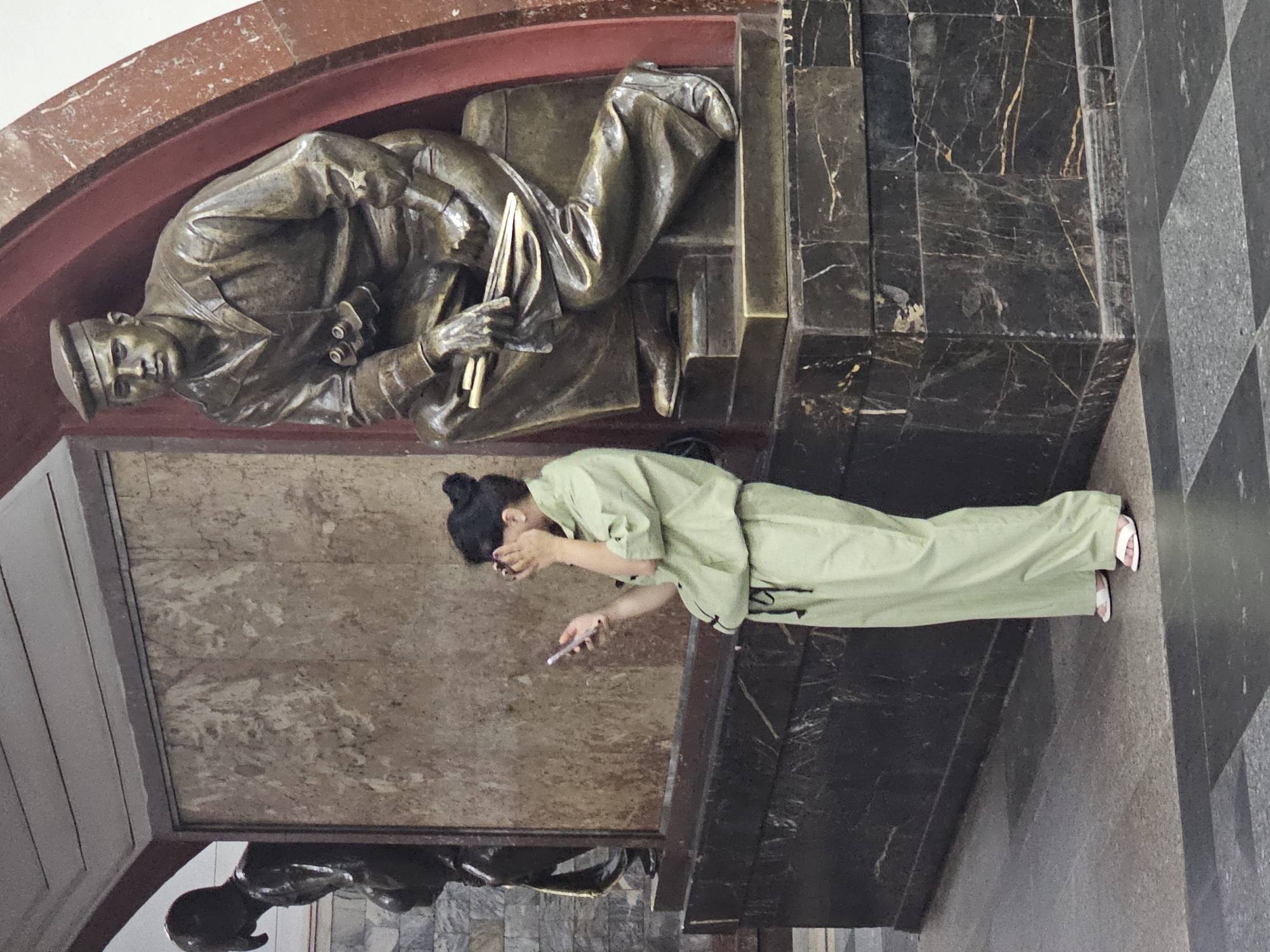 A woman in a light green jumpsuit and white sandals is standing in front of a large, dark marble pedestal that supports a bronze statue of a man in a sailor's uniform. The woman is holding a smartphone in her right hand and has her left hand covering her face. She is looking down at her phone. The statue is of a seated man wearing a sailor's cap and jacket, with binoculars around his neck. He is holding some rolled-up documents. Behind the woman and to her left is another dark bronze statue of a figure from the waist up, with its head bent forward. The background consists of a textured beige wall and an arched red structure. The floor is a patterned mosaic of dark and light gray squares. The scene appears to be set in a public indoor space, possibly a subway station or museum, in an unknown city.