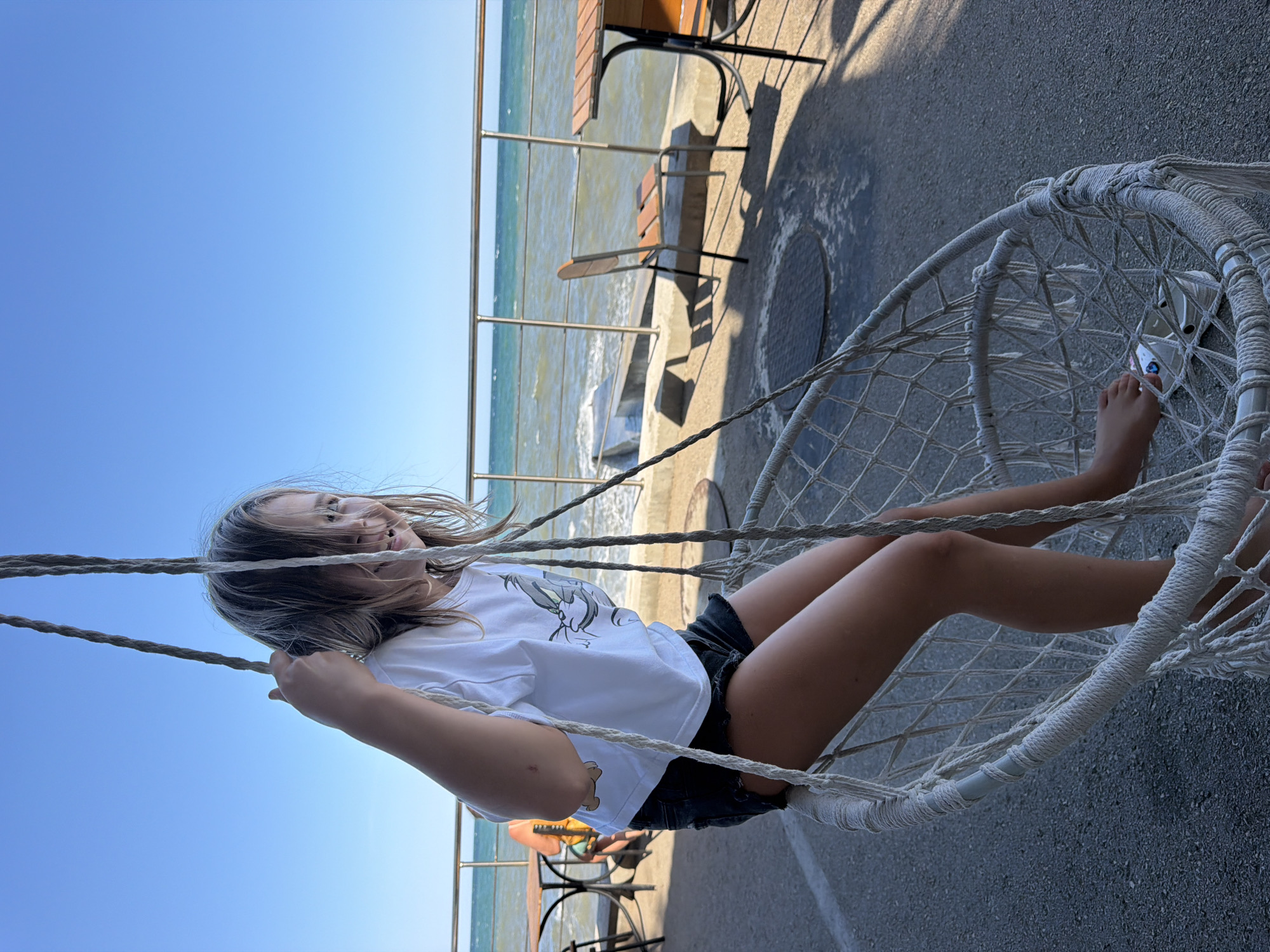 A young girl with blonde hair is sitting in a hanging swing chair. She is wearing a white t-shirt with a cartoon character and black shorts. The girl is looking to her right and smiling. In the background, the sea is visible, with a railing separating the promenade from the water. Several tables and chairs are set up outdoors, suggesting a cafe or restaurant. The setting appears to be a seaside promenade in Partenit, Ukraine.