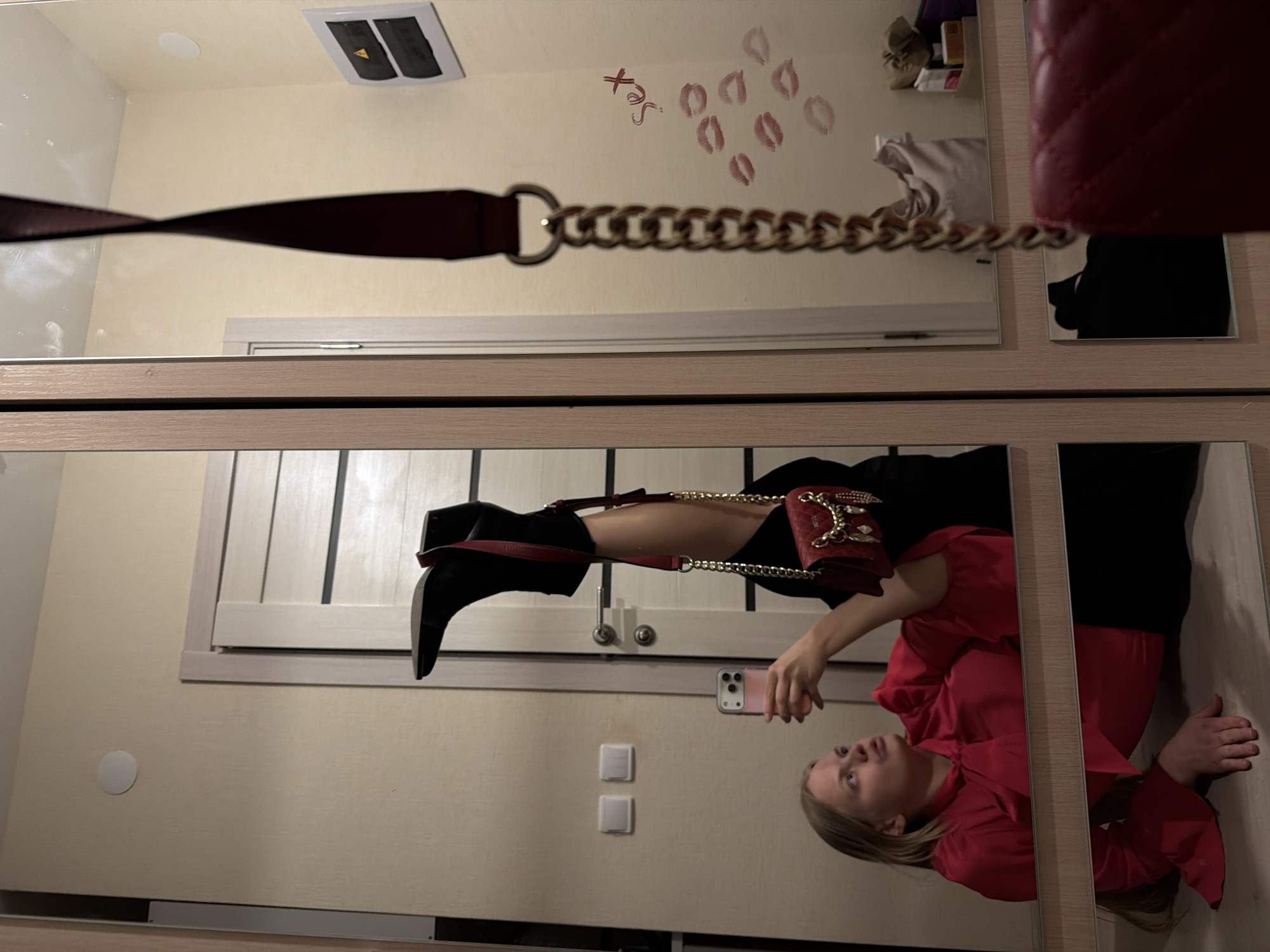 The image displays a mirror selfie taken in an indoor setting, likely in Izhevsk, Russia. The reflection in the mirror captures a young adult woman posing.
**Subjects:**
The primary subject is a woman with blonde hair, visible in the reflection. She is dressed in a vibrant red long-sleeved top with a tie neck and dark trousers or a skirt. She wears a dark, pointed-toe boot on one raised leg. A red quilted handbag with a gold chain strap hangs across her body. In her right hand, she holds a light-colored smartphone, aimed towards the mirror.
**Scene and Setting:**
The setting appears to be a room, possibly a bedroom or dressing area, indicated by a large wardrobe with light-colored wooden frames and mirrored panels. The walls are a light cream or beige color.
**Activity or Event:**
The woman is actively taking a photograph of herself in the mirror, commonly known as a mirror selfie, or documenting her outfit of the day (OOTD).
**Notable Details:**
The wall reflected in the mirror features several distinct red lipstick kiss prints arranged in rows, along with the word "Sex" written in red lipstick. On the ceiling, a white electrical panel with yellow warning labels and a circular white light fixture are visible. White light switches are present on the wall near the woman's reflection. The time of day is not discernible, but the room is adequately lit, likely by artificial light. The woman's expression is neutral as she focuses on her phone and reflection.
