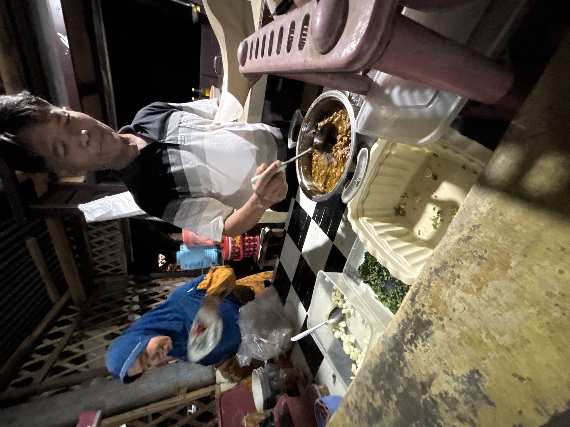 In the city of Banyakprodo, Indonesia, a man in a striped polo shirt is serving food from a pot on a checkered counter. He uses a ladle to scoop a brown stew with meat into a bowl. To his left, a woman wearing a blue hijab and an orange patterned top also appears to be involved in food preparation, holding a plate. The counter is laden with various containers of ingredients, including chopped white pieces, possibly garlic, and chopped green herbs. The setting appears to be an outdoor food stall at night, with wooden structures and ambient lighting.