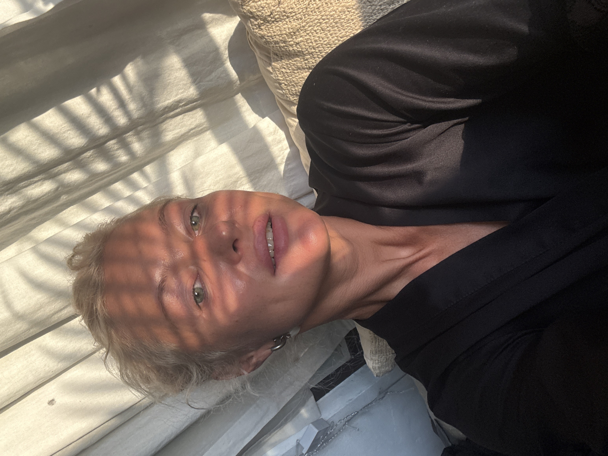 A close-up, horizontal shot captures a person lying down, bathed in dappled sunlight. The subject, with blonde hair and green eyes, looks directly at the camera, their face illuminated by the light and shadow patterns of what appears to be palm fronds. They are wearing a black garment. The background consists of light-colored fabric, possibly bedding or a curtain, with a textured pillow partially visible to the left. The sunlight creates strong contrasts on the subject's skin, highlighting their features and the texture of their face. The overall mood is relaxed and contemplative, with the interplay of light and shadow adding a dramatic effect. No visible text is present in the image.
