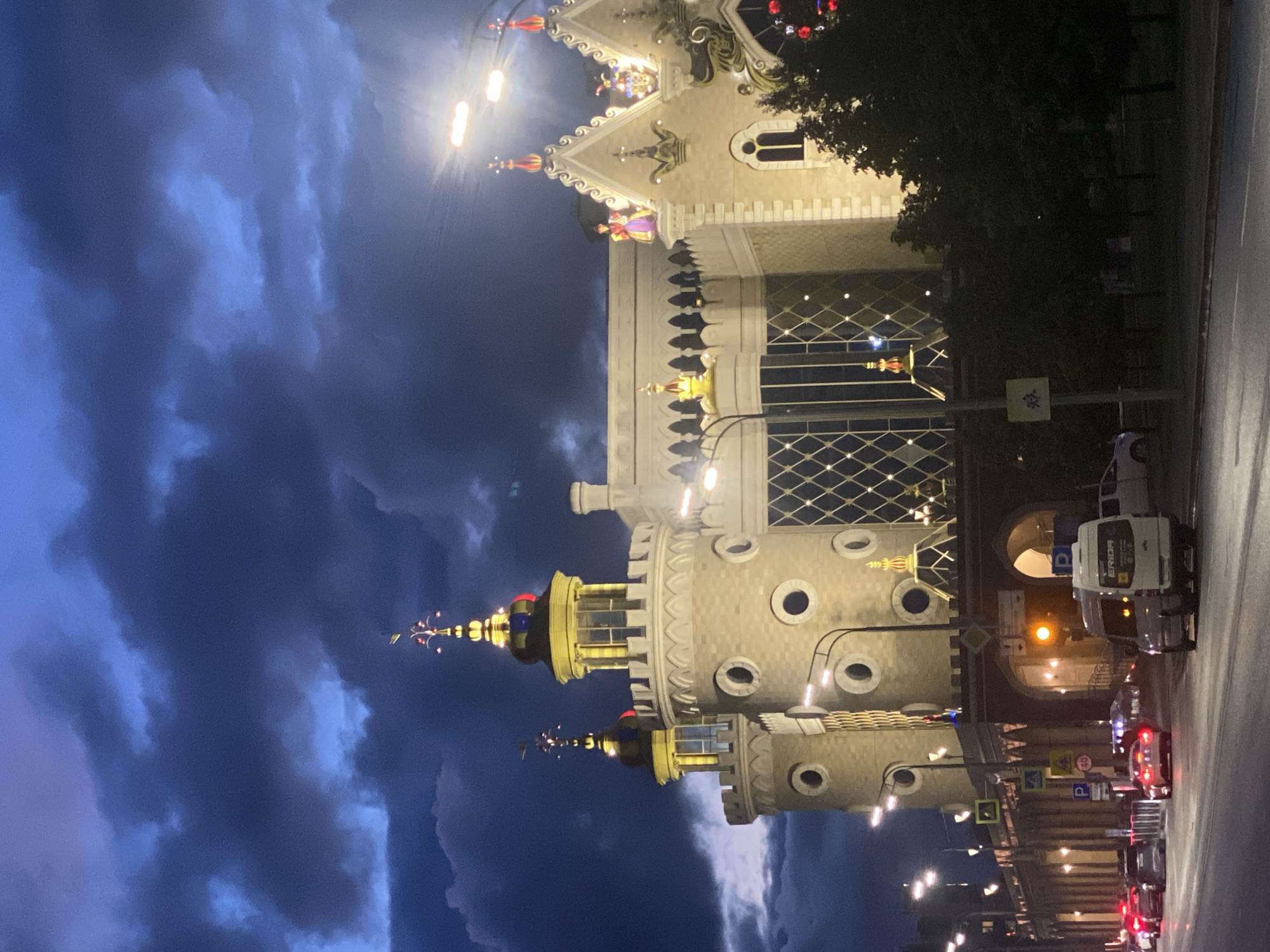 The image, taken at night in Kazan, Russia, showcases the distinctive architecture of the Kazan Puppet Theatre 'Ekiyat' under a dramatic, cloudy sky. The building, illuminated by numerous lights, features an intricate, fairytale-like design with multiple towers, pointed gables, and decorative spires topped with gold and red elements. Several round windows are visible on the towers, and the roofline is adorned with ornate detailing and what appear to be figures or festive decorations. A dense tree partially obscures the right side of the main building.
In the foreground, a street runs parallel to the theatre. Streetlights cast a warm glow on the road, where several vehicles, including cars with illuminated headlights and taillights, are present. A white van, prominently displaying the text "ERIDA," is parked or driving on the right side of the road. Various road signs, including blue "P" parking signs, are visible along the street. The sky is a deep blue and purple, filled with textured, heavy clouds, indicating a late evening or night scene. No explicit human activity other than evening traffic is discernible. The overall impression is one of a magical, brightly lit landmark set against a moody, twilight backdrop.