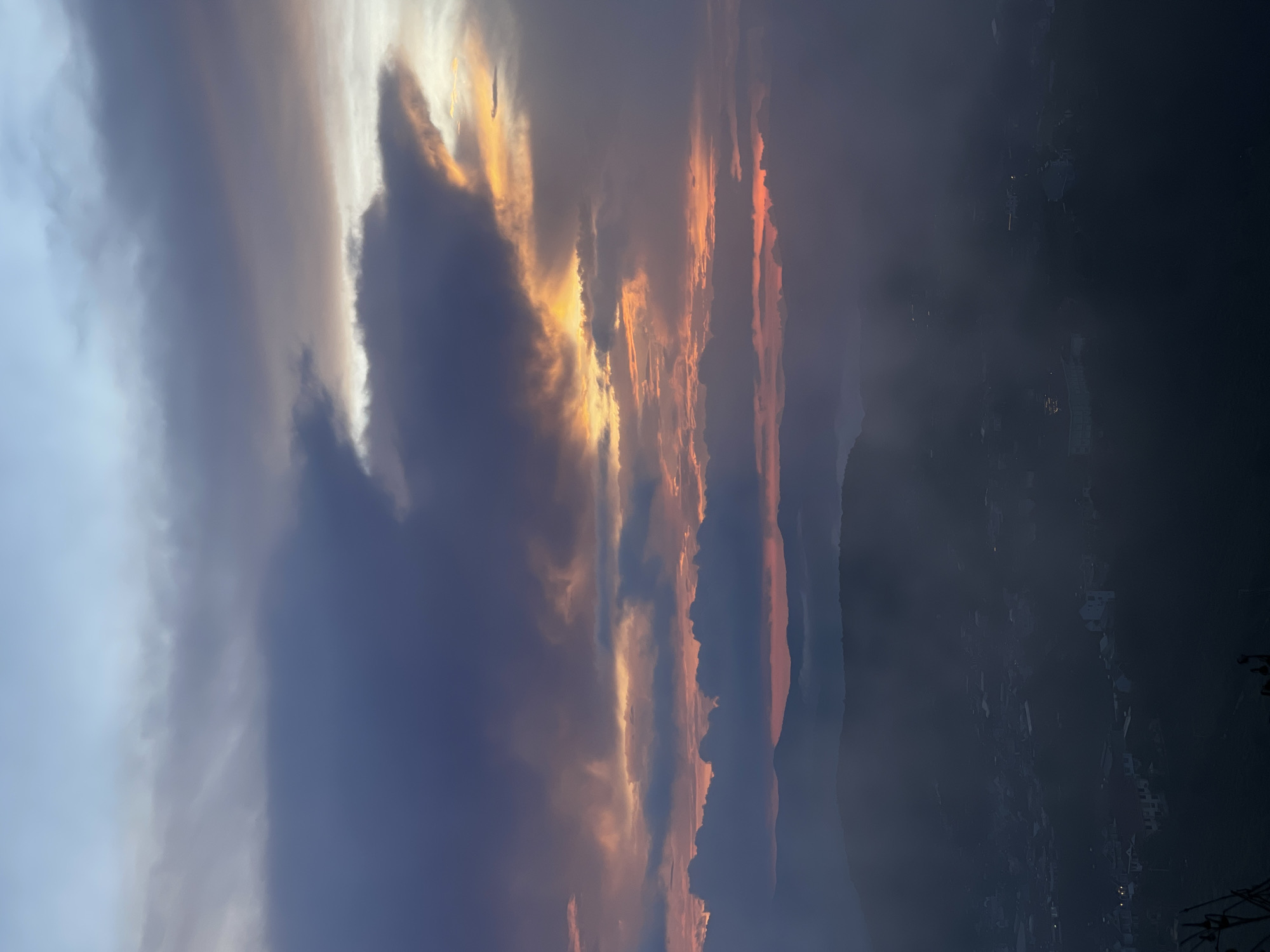 The image shows a dusky sky over Trường Thọ, Vietnam. Thick, dark clouds fill much of the frame, some streaked with orange and gold hues from the setting sun. Lower in the frame, a darkened cityscape with scattered lights is faintly visible. Mountains are silhouetted in the mid-ground. The image is vertically oriented.