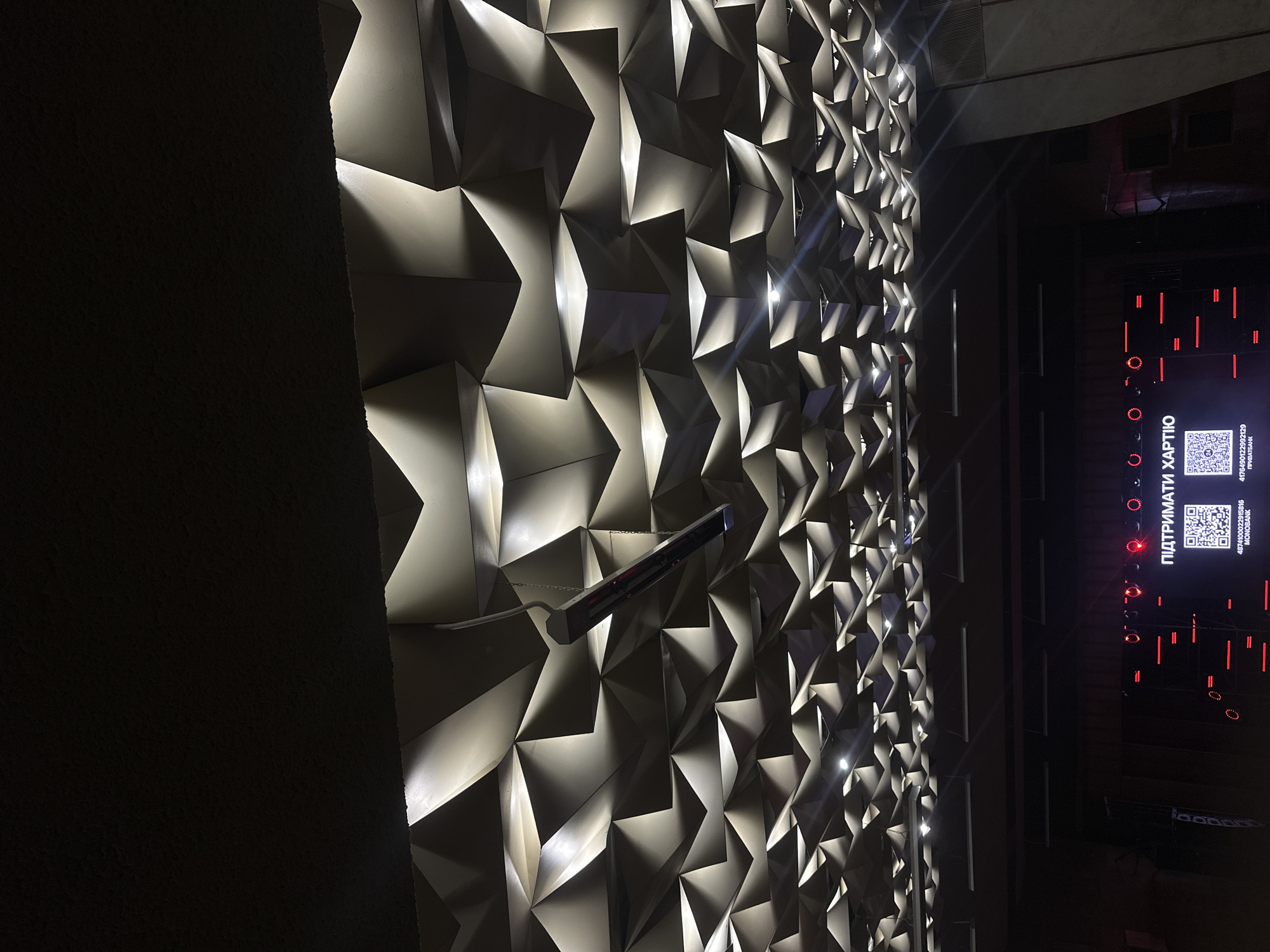 The image shows the interior of a modern event space or concert hall, likely located in Ternopil, Ukraine. The most striking feature is the ceiling, which is covered in a complex, geometric, three-dimensional design with integrated lighting. This creates a dramatic and visually engaging effect.
Below the ceiling, a stage area is visible. A large screen displays the Ukrainian text "ПІДТРИМАТИ ХАРТІЮ" (Support the Charter). There are also two QR codes displayed on the screen, one labeled "MONOBAМК" and the other "ПРИВАТБАНК", indicating ways to donate or support the cause. The stage is decorated with red neon lights and red circular lights. A single, long, dark-colored heater or projector is suspended from the ceiling by chains, hanging centrally in the upper part of the image.
The overall environment appears to be a well-lit, modern venue designed for events, possibly a theater, concert hall, or conference center. There are no people visible in the frame.
