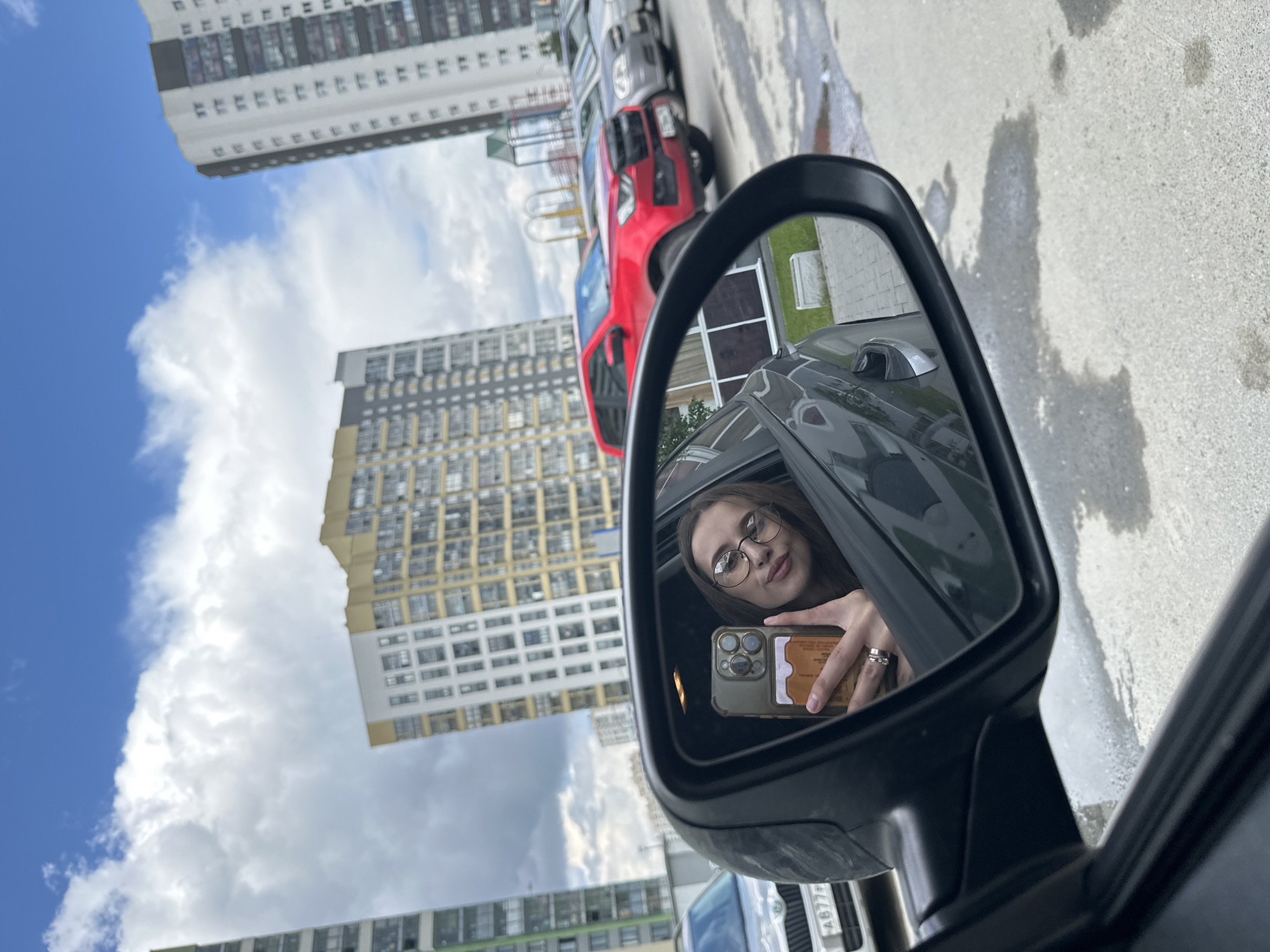 A detailed image captures a selfie reflection in a car's passenger-side mirror within a Yekaterinburg, Russia, urban environment.
The central subject is a young woman, visible in the mirror's reflection, seated inside a dark-colored vehicle. She possesses long, dark hair and wears round-framed glasses. Her right hand is raised, holding an iPhone with a multi-lens camera module, encased in a clear case with a brown card (possibly an ID or credit card) inserted into a slot on the back. A ring is visible on her right ring finger. She exhibits a subtle smile, indicating she is taking a self-portrait.
Outside the car, the immediate foreground shows wet, grey asphalt or concrete pavement with several distinct puddles or damp patches. Parked cars line the street. Directly adjacent to the vehicle from which the photo is taken is a silver Skoda Octavia, partially visible, bearing a Russian license plate "A877PE 196", where '196' is the region code for Sverdlovsk Oblast, encompassing Yekaterinburg. Further down the street, a prominent red SUV (likely a Haval model) is parked, followed by several other grey sedans. A multi-colored children's playground structure is visible behind the red SUV.
The background features several modern, multi-story residential buildings. One on the far left is green and grey, while a large structure in the center is primarily yellow and white with grey accents. Another tall, white and grey building with dark windows stands on the far right. The sky overhead is partially cloudy with significant patches of bright blue. Some green foliage and a white planter box are visible at the base of the buildings.