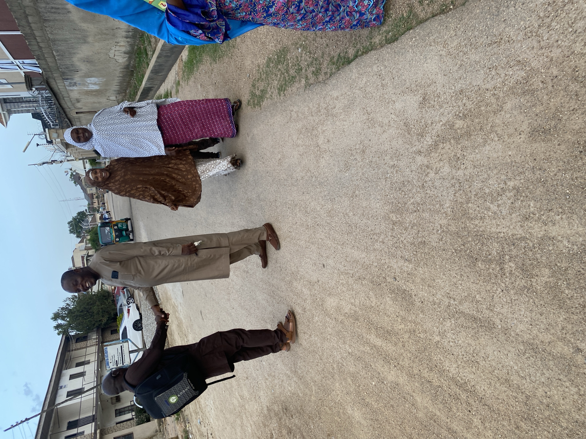 Four individuals are captured in this outdoor photograph, seemingly engaging in a social interaction. The image is oriented such that the ground appears to be on the right side and the sky on the left.
In the foreground, two men are shaking hands. One man, wearing a brown robe and carrying a black bag, is on the left. The other man, dressed in a light brown robe, is on the right, smiling broadly.
Further in the background, two women are standing. The woman on the left is wearing a white patterned top and a purple skirt, with a headscarf. The woman to her right is in a brown headscarf and dress.
The scene appears to be a street in Jos, Nigeria. Buildings and a three-wheeled vehicle (keke napep) are visible in the background. The weather seems overcast, suggesting it might be early morning or late afternoon. The overall mood of the image appears positive, with the smiling faces of the individuals. The ground is a rough, sandy texture.