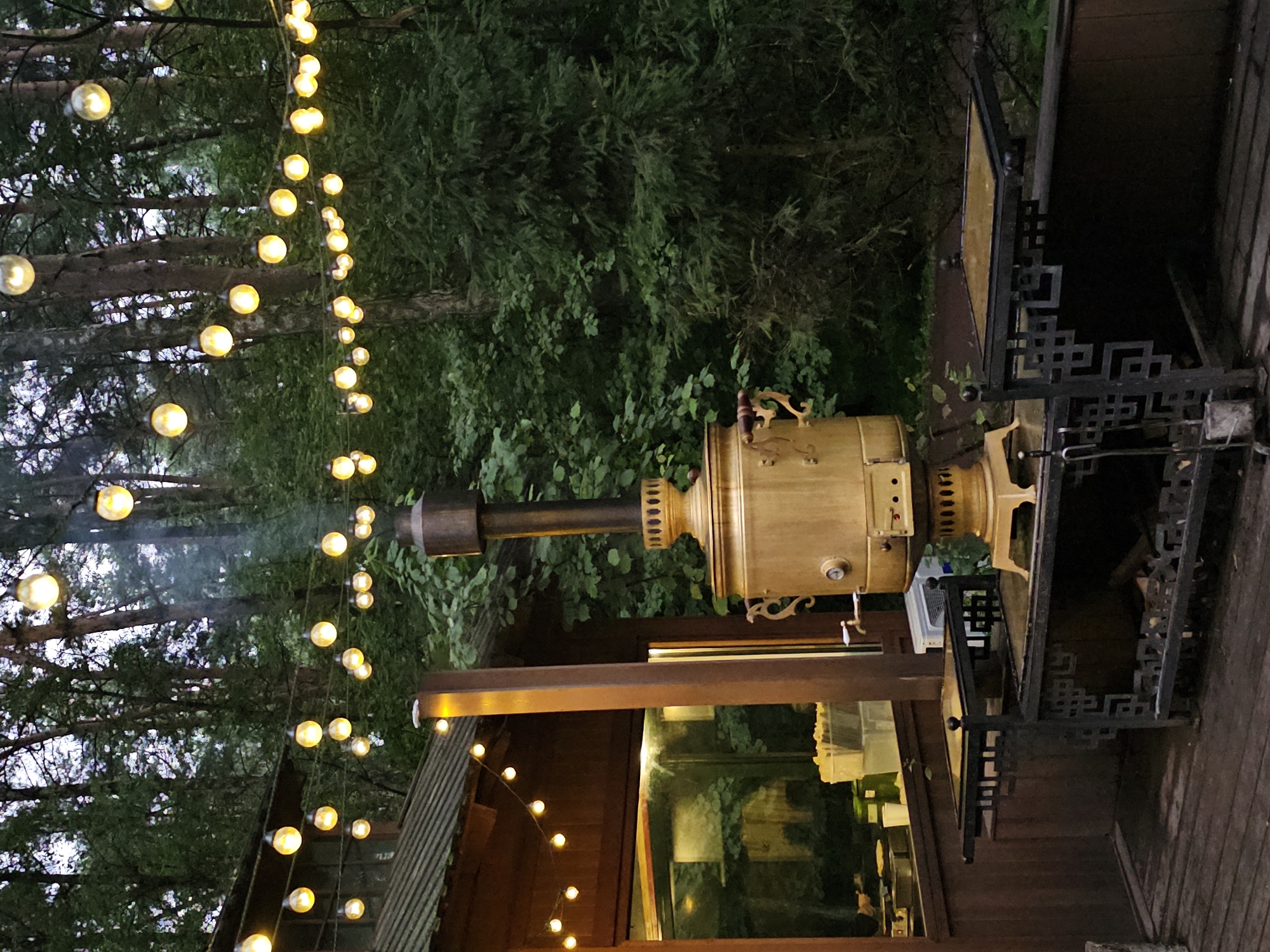 The image depicts an outdoor scene at dusk, featuring a wooden structure with a porch and a string of lights overhead. The focal point is a large, traditional brass samovar emitting a thin stream of smoke, placed on a metal stand. Behind the samovar, a dense forest with tall trees creates a natural backdrop. The lighting suggests an evening setting, with the string lights casting a warm glow. The overall atmosphere is rustic and serene, with elements of both human-made and natural environments. The location is identified as Nemchinovo, Russia.