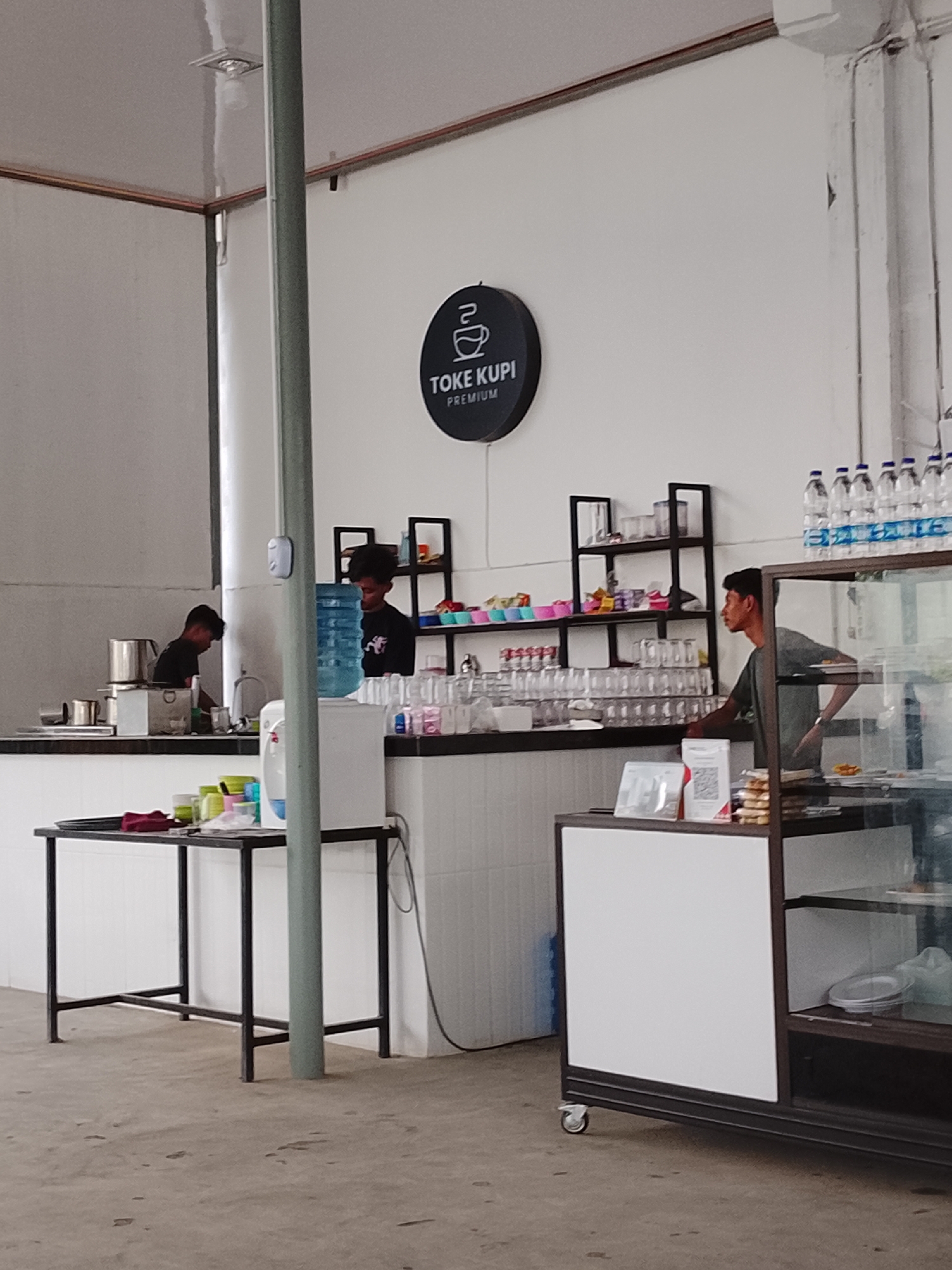 The image shows the interior of a cafe named "Toke Kupi Premium" in Lhokseumawe, Indonesia. Three men are present, two behind the counter and one in front. The counter is lined with glassware and various supplies. A black sign with the cafe's logo is mounted on the wall above the counter. A metal table with a black frame holds cups and other items. Bottled water is stacked above the display case to the right of the frame. The floor is concrete.