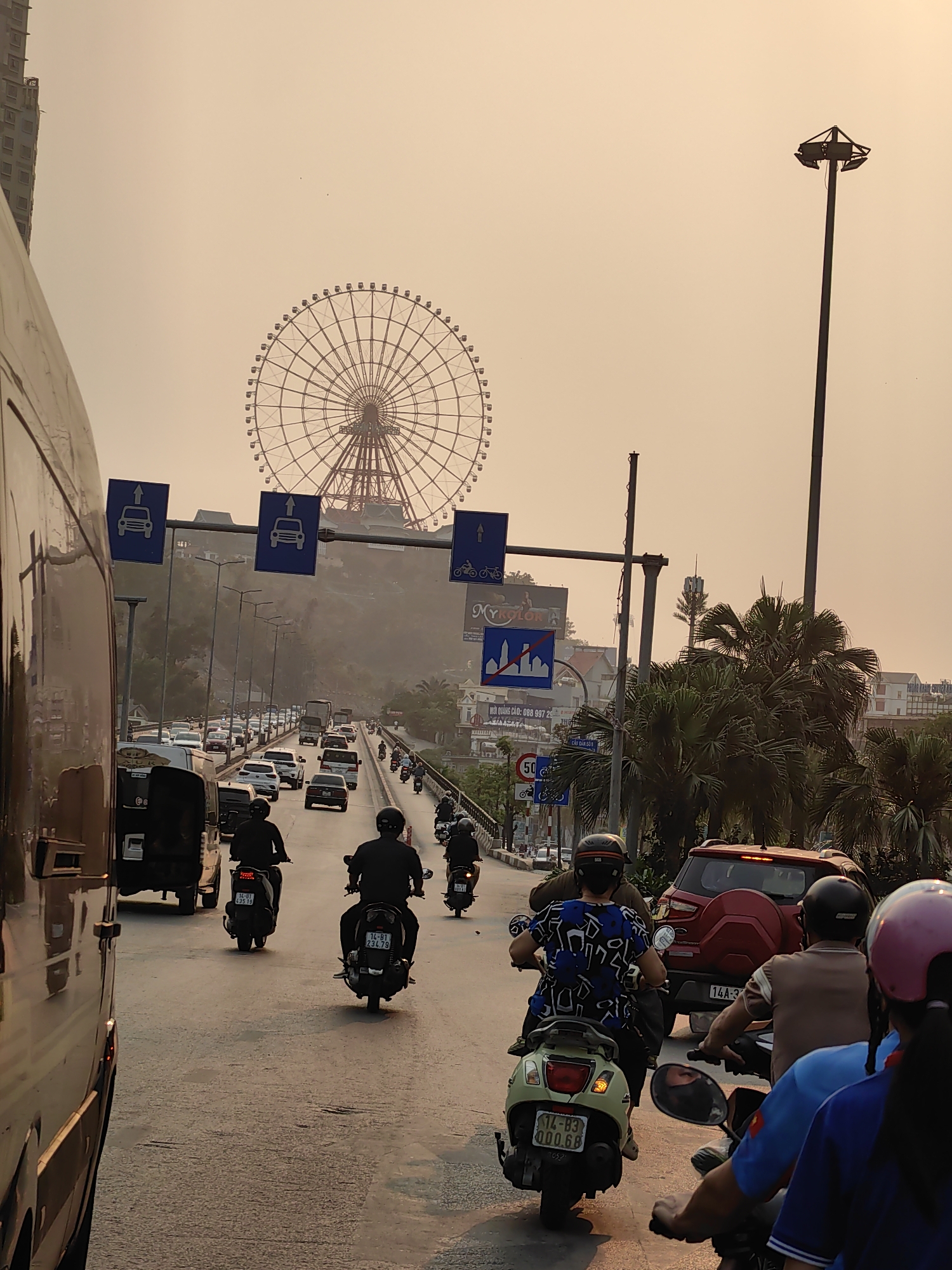 The image captures a busy street scene in Hong Gai Ward, Vietnam. Heavy traffic consisting of cars, motorbikes, and vans is visible, creating a congested atmosphere. Several individuals on motorbikes are heading in the same direction as the camera. A large Ferris wheel looms in the background. The air appears hazy, casting a warm light over the scene. Street signs and a cell tower are also present, along with palm trees and other vegetation. A red SUV is partially visible among the traffic. The road appears to be a bridge or overpass, elevated above the surrounding terrain.