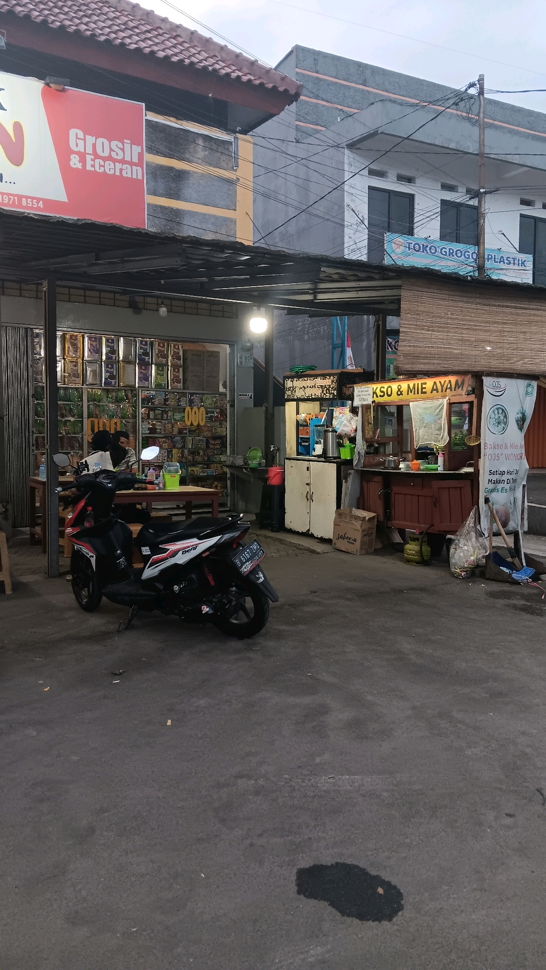 The image depicts a street scene in Depok, Indonesia, at dusk. In the foreground, a black and red motorcycle is parked on an asphalt surface with some dark stains. To the left, a covered outdoor dining area with wooden tables and chairs is visible. Behind this, a store with numerous shelves stocked with goods is illuminated from within. Further back and to the right, a food stall selling "Bakso & Mie Ayam" is set up. The stall has a wooden counter, a variety of containers, and signage. A person is partially visible behind the counter. Above the food stall, a blue banner with white text reads "TOKO GROGO PLASTIK". The background consists of multi-story buildings with windows and electrical wires. The lighting suggests it is late afternoon or early evening.