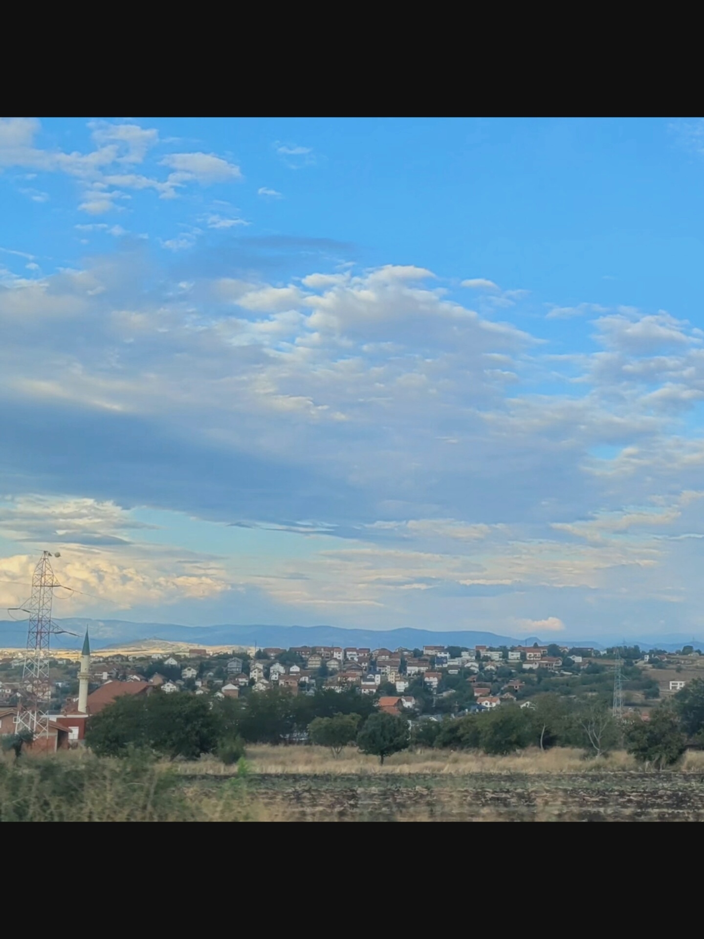 The image displays a vast, open sky filled with scattered white and gray clouds against a vibrant blue backdrop. Beneath the sky, a sprawling residential area is visible, characterized by numerous houses with red-tiled roofs, clustered together on undulating terrain. A prominent minaret, likely belonging to a mosque, stands tall amidst the buildings, alongside a tall, lattice-like power transmission tower. The foreground consists of a field of dry grass and vegetation, suggesting a rural or semi-rural environment. The overall scene conveys a sense of a settled community under a dynamic sky.