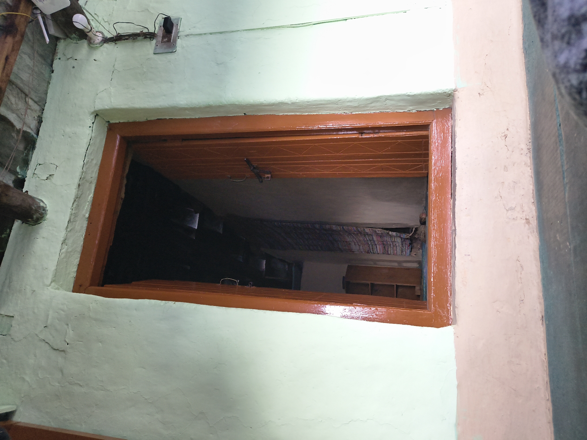 A close-up view of a window frame painted in glossy brown against a light greenish-cream wall. The frame is rectangular, and the view through the window shows a cluttered interior space with dark areas and items suggesting a residential setting. The wall has a rough texture, with some cracks and uneven surfaces. The window latch is visible on the upper part of the frame. Wires and an electrical outlet are visible in the upper left corner of the image. The location is identified as Haini, India, but no details confirm this beyond the description of the scene. The description is based on visual aspects of the image alone.