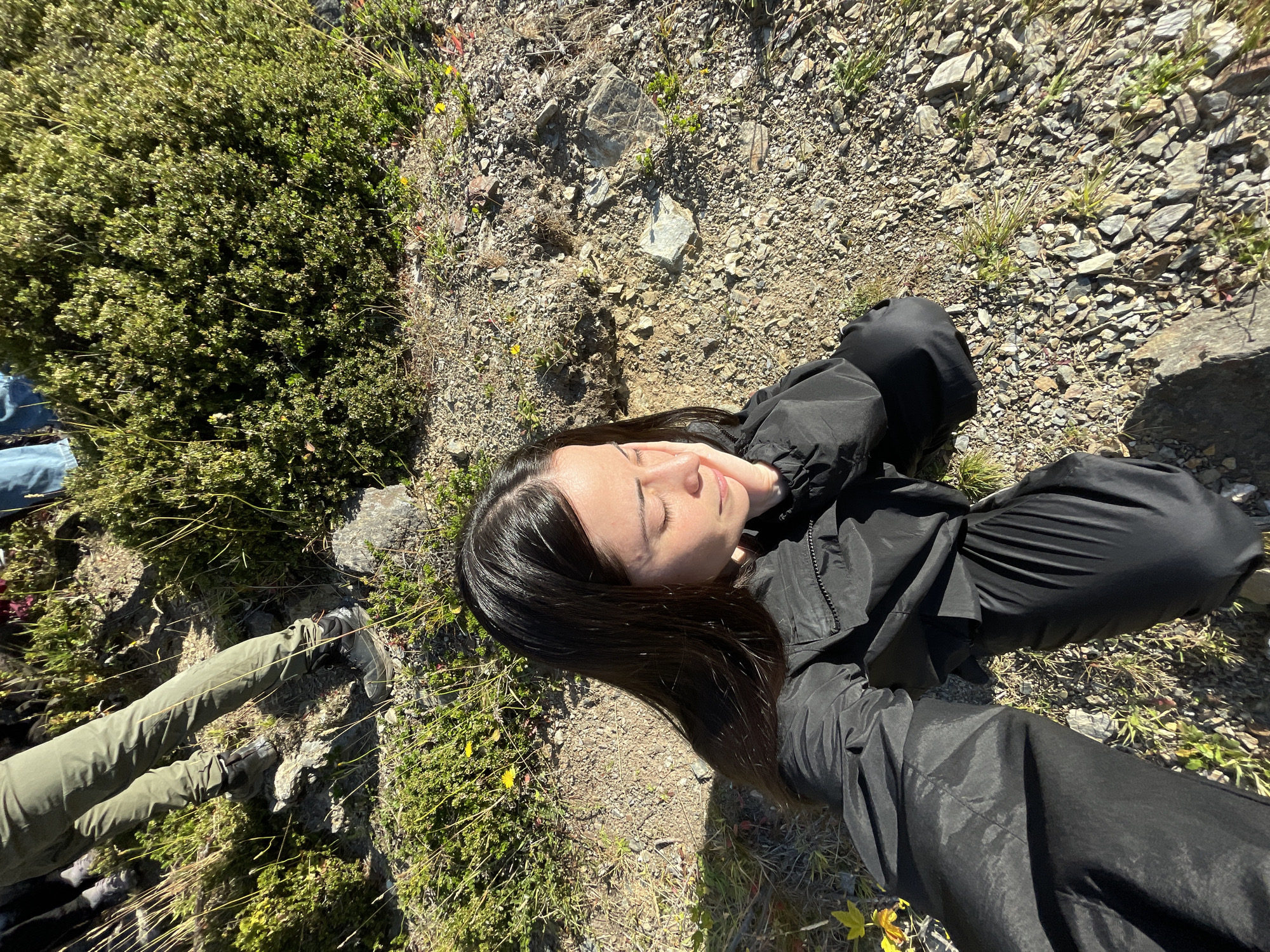 A young woman is lying on a rocky, grassy hillside. She has long, dark hair and is wearing a black jacket and pants. Her eyes are closed, and she appears to be resting. To her left is a patch of low-lying green vegetation, and to her right, the ground is covered in small rocks and sparse grass. In the far left of the frame, two legs clad in green pants are visible. The location is possibly a natural environment near Ushuaia, Argentina, based on the type of vegetation and terrain commonly found in the area.