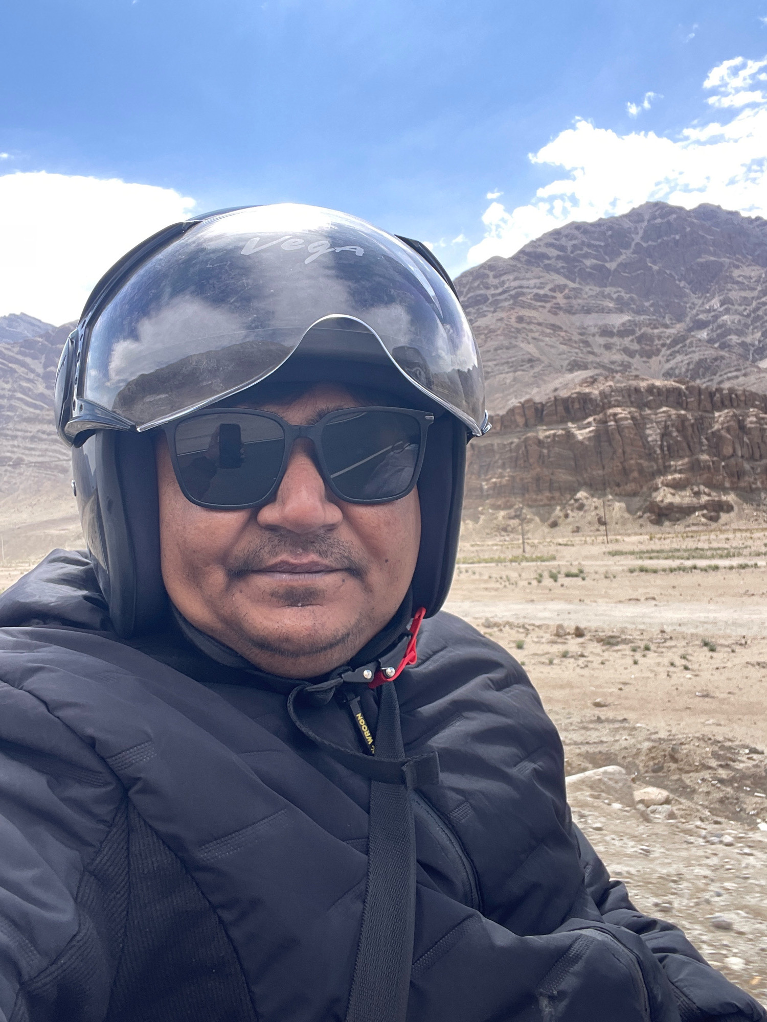 A man wearing a black helmet and sunglasses is captured in a close-up selfie, with a mountainous landscape under a cloudy blue sky in the background. He is wearing a black puffer jacket. The terrain appears arid and rocky. The man's face is the central focus of the image, with the helmet and jacket framing him. The reflection in his sunglasses shows a hand holding a smartphone, indicating he is taking the selfie. The location is Upshi, India.