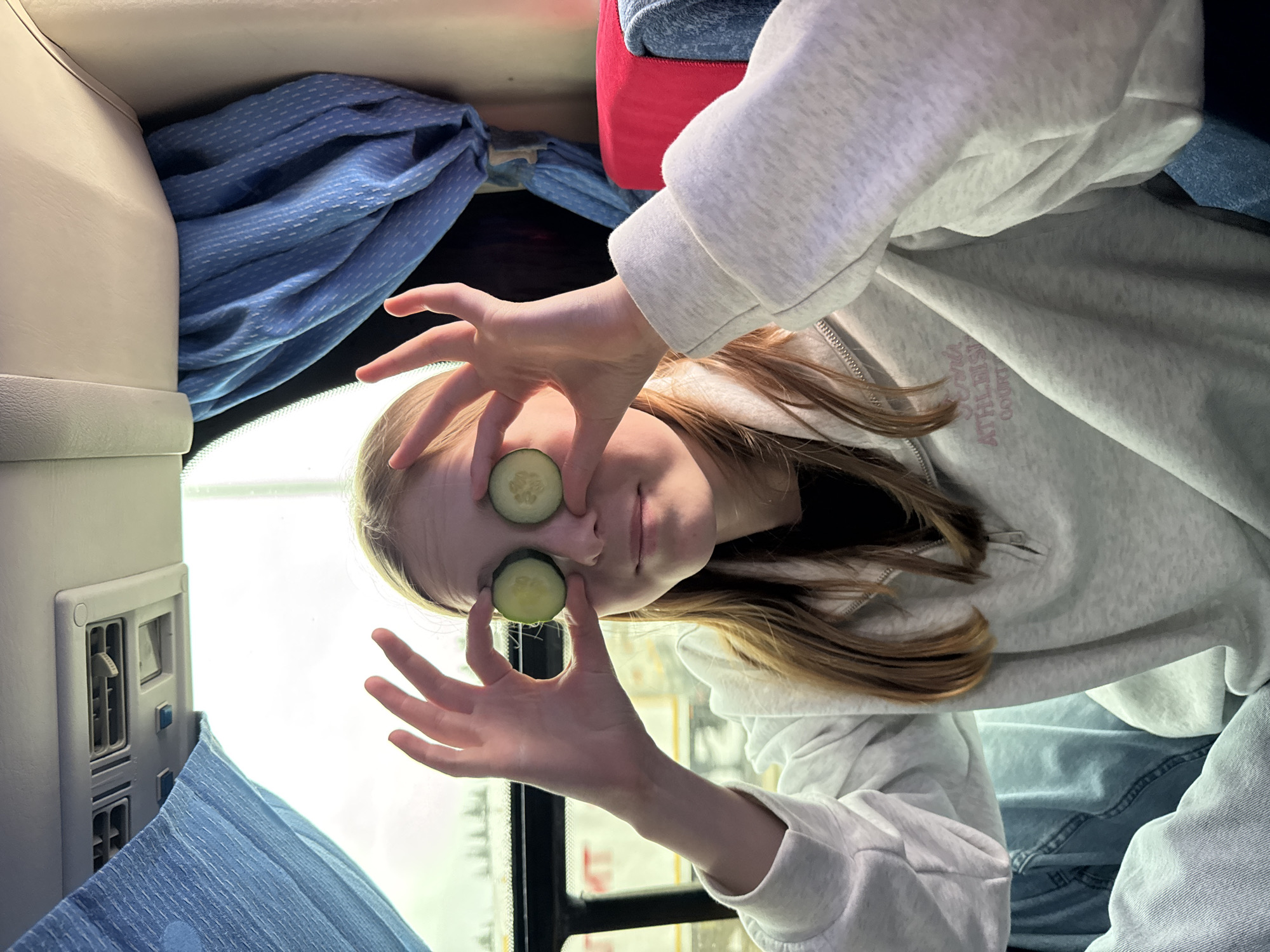 A woman with fair skin and light brown hair is holding two slices of cucumber over her eyes. She is wearing a light gray hooded sweatshirt. The background suggests she is inside a vehicle, possibly a bus or train, with a window, blue curtains, and overhead storage compartments. The location of Yanychi, Russia is not discernible from the visual information provided.