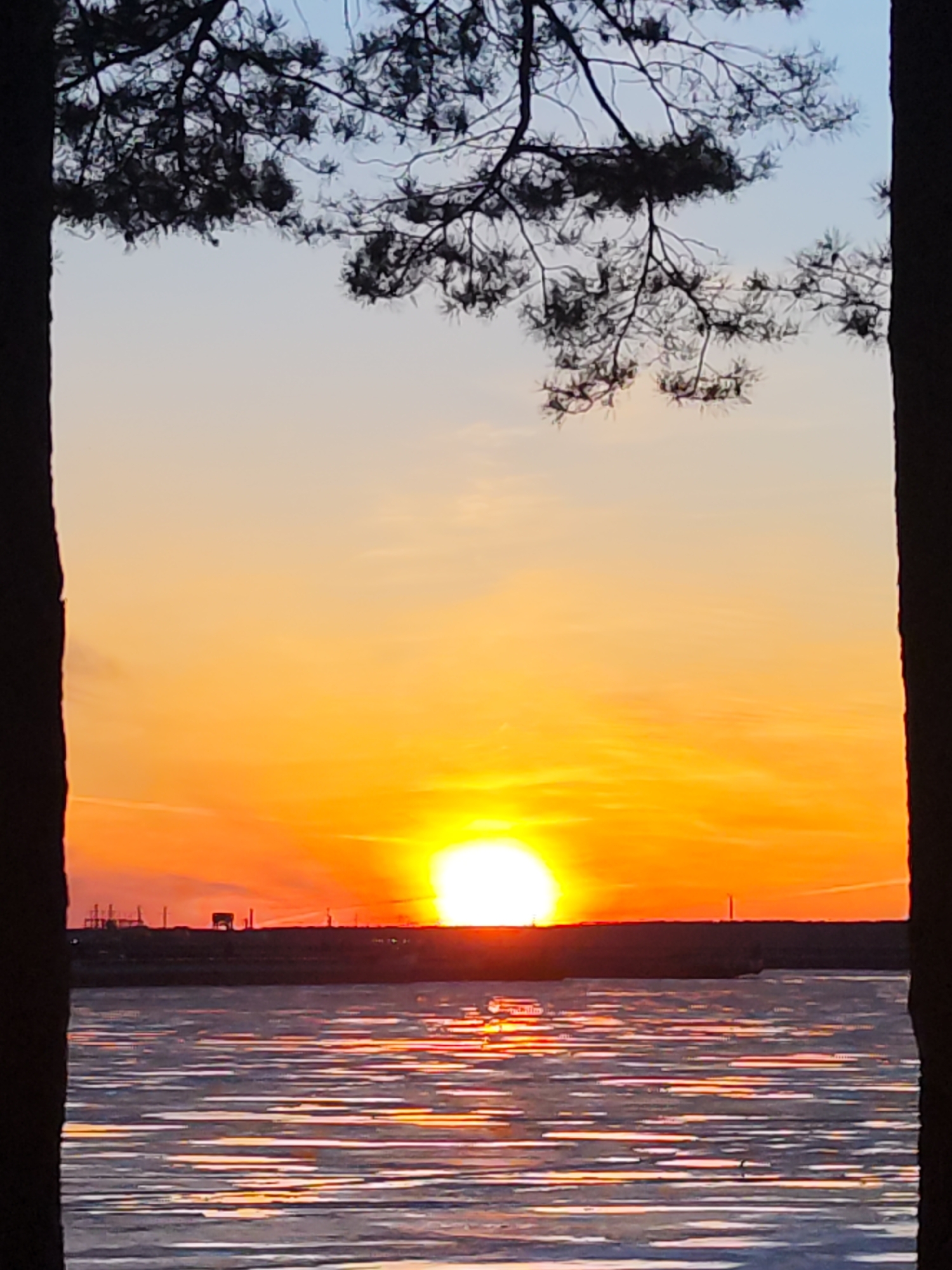 The image depicts a sunset over a body of water. The sun is low on the horizon, emitting a bright, intense light, with the sky transitioning from orange near the horizon to a lighter blue higher up. The water reflects the sunlight in streaks and bands across its surface. The silhouette of trees, branches, and foliage frames the scene at the top and sides of the image. In the distance, on the horizon line, are indistinct structures and possibly industrial elements.
Based on the description of the location, the most distinct features available would be the sunset and the horizon line. Without additional geographical features or distinguishing architectural elements in the provided image, confirming the specific location within Chaikovsky, Russia, is not possible with certainty.