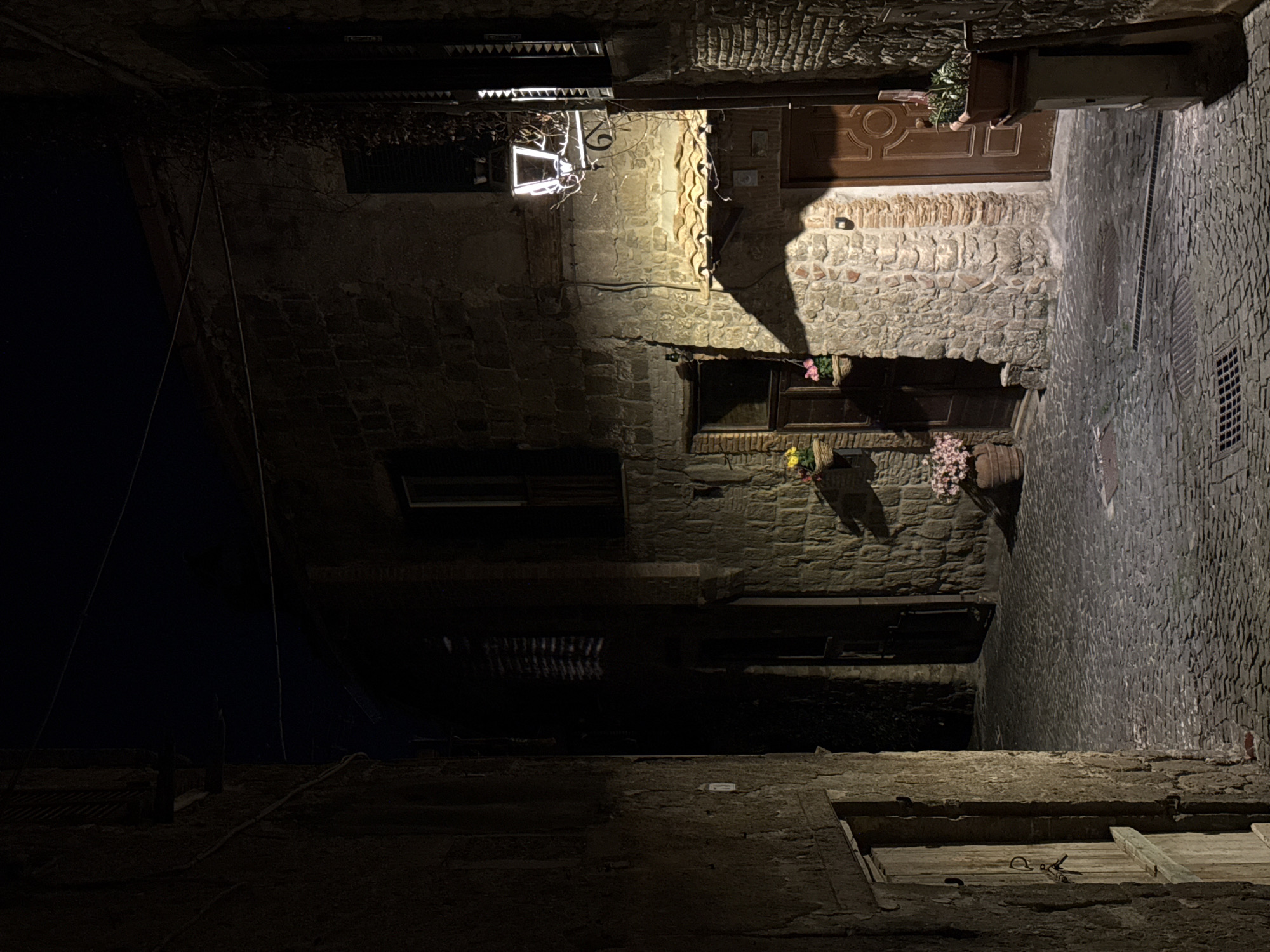 The image depicts a narrow, dimly lit street in Castelnuovo di Porto, Italy, at night. The view is looking upwards.
The street is paved with cobblestones. On the right side is a wall constructed of irregularly shaped stones. A drain is set into the pavement.
Buildings line the street on both sides. The architecture is old, with stone walls and small windows. Flower boxes are visible in some windows. A wooden door is visible on the upper-right. An ornate light fixture illuminates the space.
The sky above is dark, suggesting it is nighttime. The overall impression is one of a quiet, historic European street after dark.