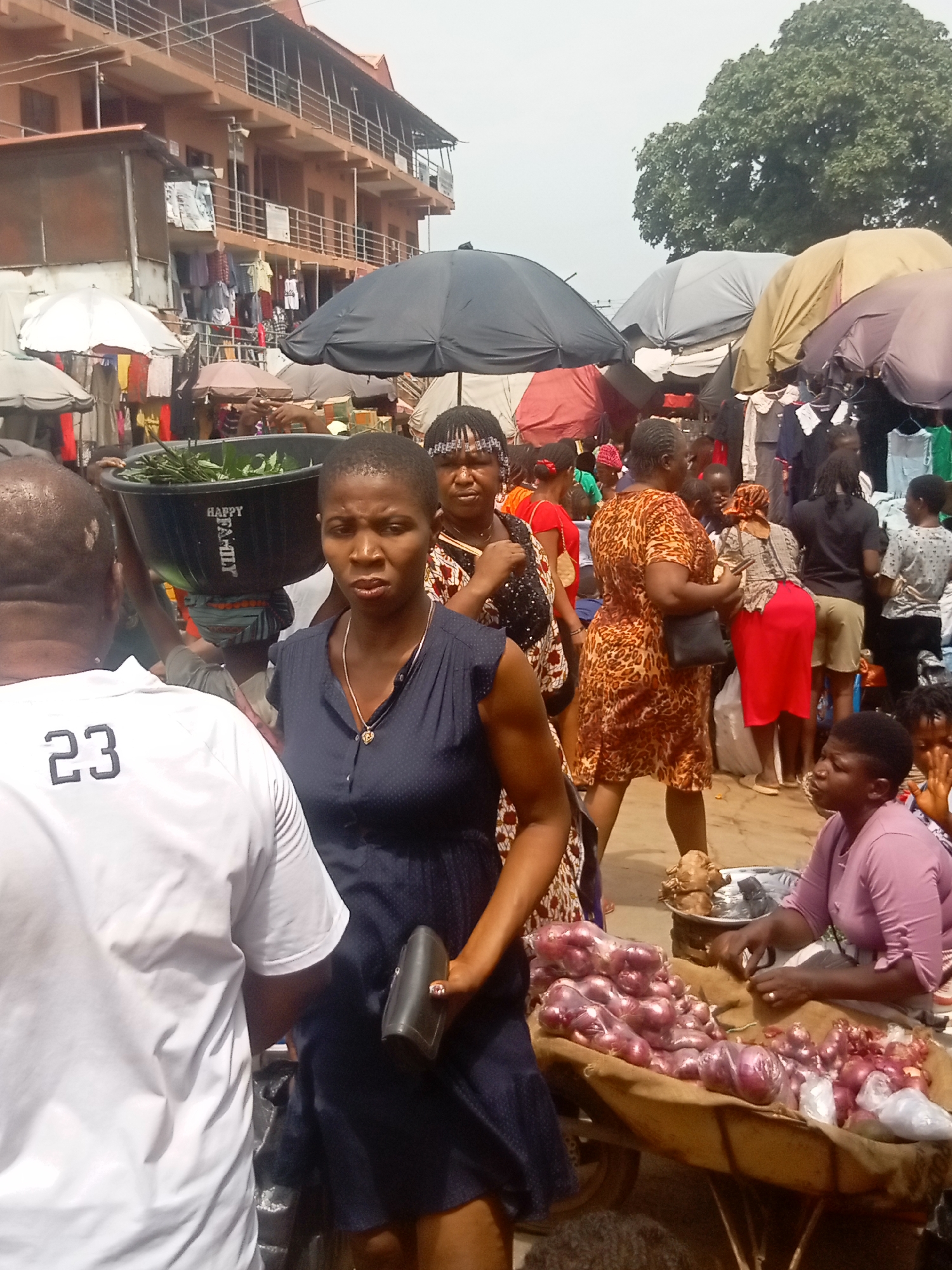The image captures a bustling market scene in Onitsha, Nigeria. In the foreground, a woman with a dark blue dress and a wallet is standing next to a man wearing a white shirt with the number 23 printed on it. Behind her, another woman has a large bowl of greens balanced on her head. To the right, a vendor sits beside a cart loaded with red onions, apparently selling them. Various other people are visible, some under umbrellas, browsing clothing and other items. Buildings are visible in the background, suggesting this is an established part of the city market.