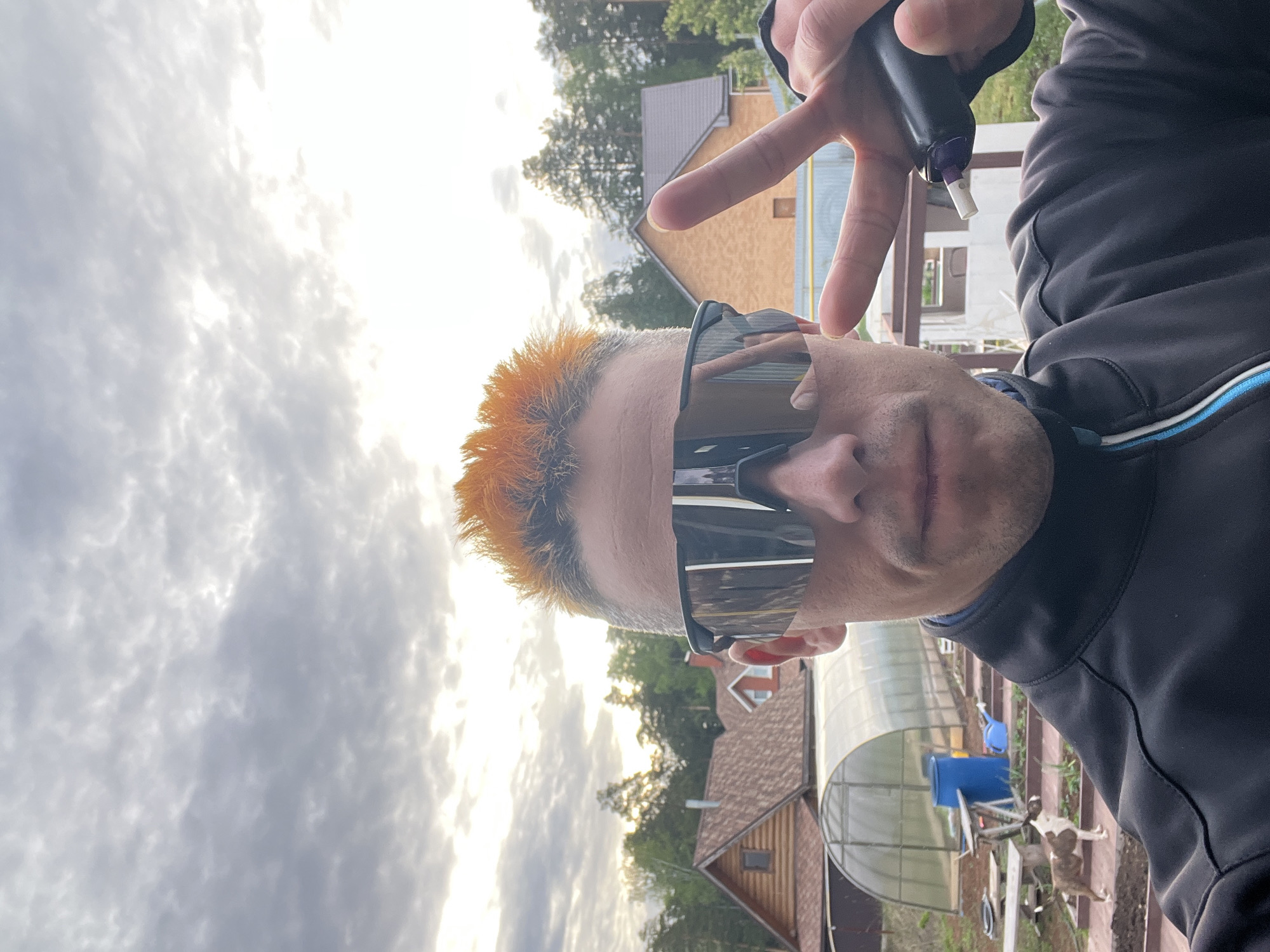 A man with orange hair and dark sunglasses is making a peace sign. He is wearing a black jacket with blue and white trim. In the background, there are buildings, trees, a greenhouse, and cloudy skies. The location is near a house with a brown roof and siding. There are blue containers and gardening equipment visible in the background. A dog is lying on the ground. The photo appears to be taken outdoors. It is difficult to confirm if the exact location is Chaikovsky, Russia, based solely on the visual information provided.