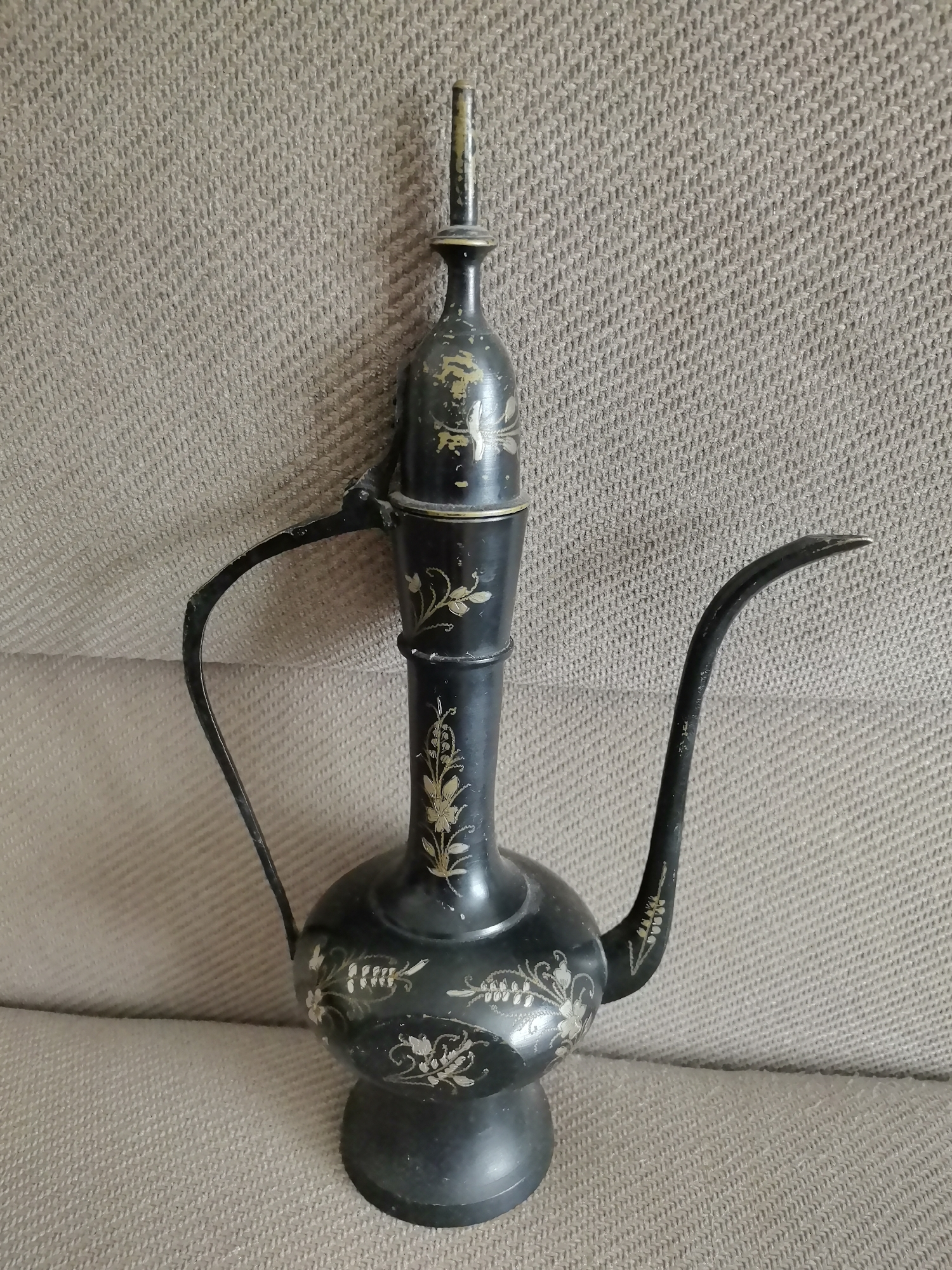 An antique, dark-colored metal ewer with ornate gold detailing sits on a textured, neutral-colored surface, possibly a sofa or upholstery. The ewer has a bulbous base, a slender neck, a curved handle, and a long, thin spout. The gold decorations appear to be floral or vine-like. The background is a uniform, textured fabric. There are no people or animals visible. The location is not specified and cannot be determined from the image.