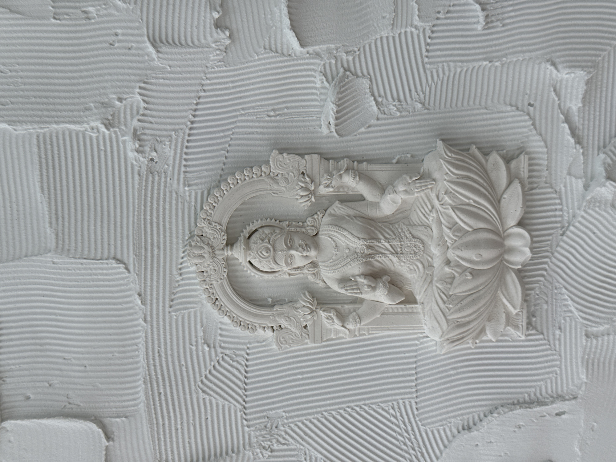 An ornate, white, bas-relief sculpture of a Hindu deity is embedded in a heavily textured white wall. The sculpture is detailed, featuring a figure reclining on a lotus flower and framed by an ornate halo. The wall's texture is created by parallel lines of white material applied in different directions, forming a cross-hatched pattern around the sculpture. The location is Sochi, Russia.