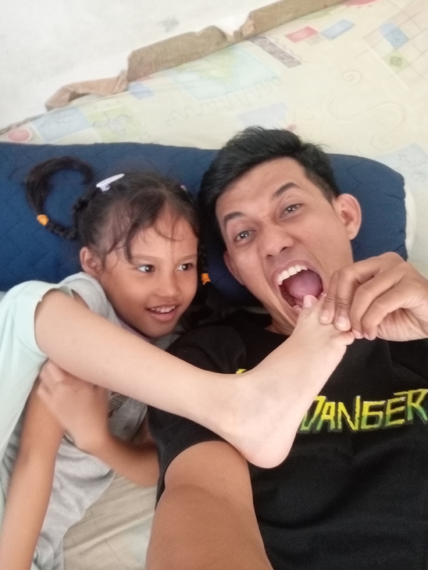 The image shows a father and daughter lying on a bed. The father, a man with short black hair, is wearing a black t-shirt with yellow and green text that reads "DANGER". He is holding his daughter's foot up to his mouth, with his mouth open as if he is about to bite it. The daughter, a young girl with brown hair in pigtails, is wearing a light blue t-shirt. She is smiling and looking towards the father. The background appears to be a bedroom with a patterned bedspread. The lighting suggests it is daytime. The emotion conveyed is playful and affectionate. This is a candid, close-up shot of a lighthearted moment between a father and his daughter.