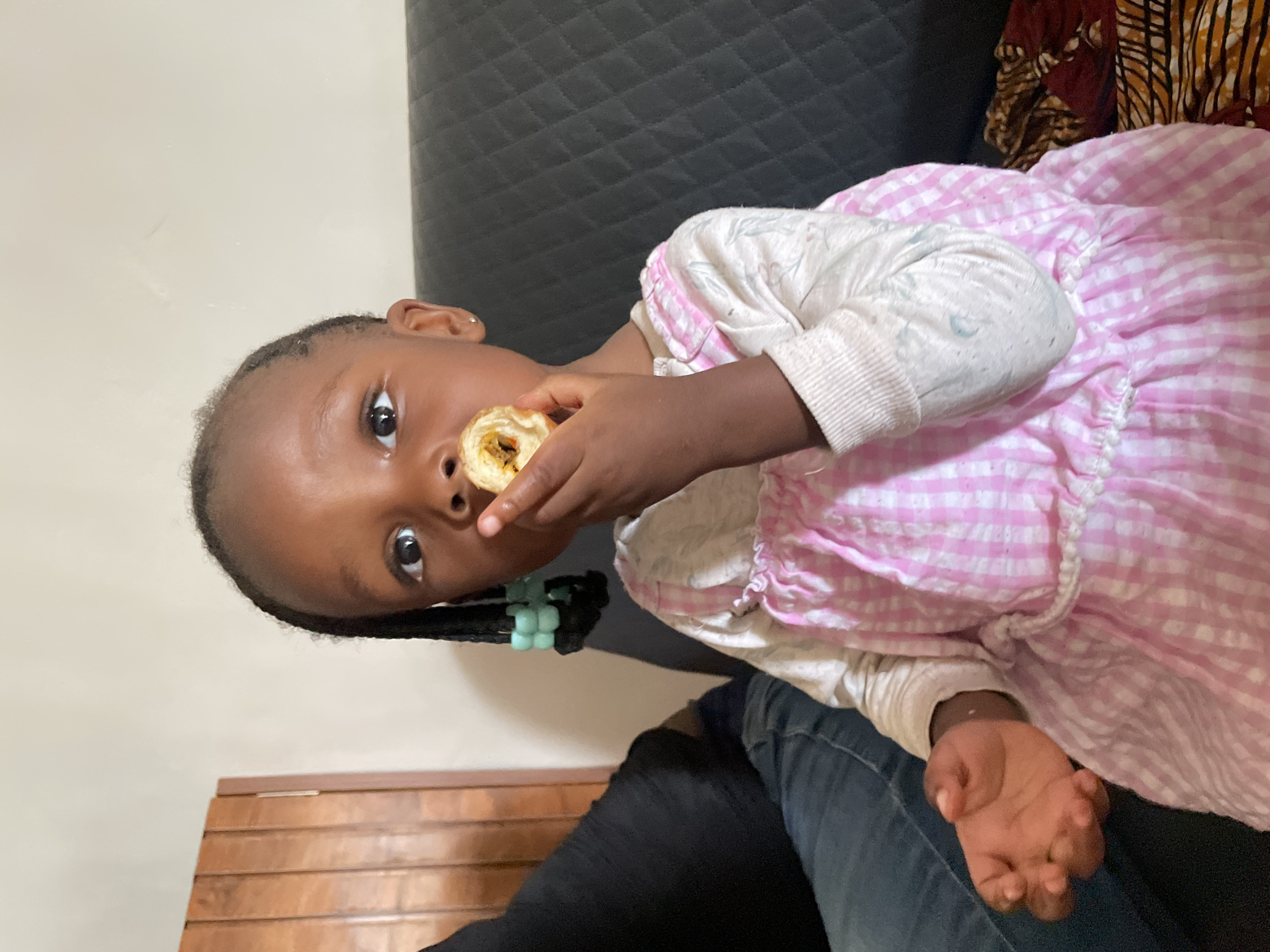 The image features a young child, likely a girl, captured indoors in Barakin Akawo, Nigeria. She has dark skin, large expressive dark eyes, and dark hair styled in braids with light blue beads. The child is wearing a white long-sleeved top beneath a pink and white gingham checkered dress, and dark blue jeans cover her legs.
She is actively eating, holding a light brown, rolled-up pastry or snack with a visible dark filling in her right hand, bringing it towards her mouth. Her gaze is directed straight at the camera with a wide-eyed, innocent, and engaged expression.
The setting appears to be a living room or common area, with the child seated on a dark, possibly quilted sofa or couch. In the background, part of a patterned fabric (possibly a cushion or throw) with warm tones of brown, orange, and black is visible to the upper right. A plain light-colored wall stretches behind the child, and a section of wooden furniture is present in the lower left corner. The ambient light suggests it is daytime. There is no visible text in the image.