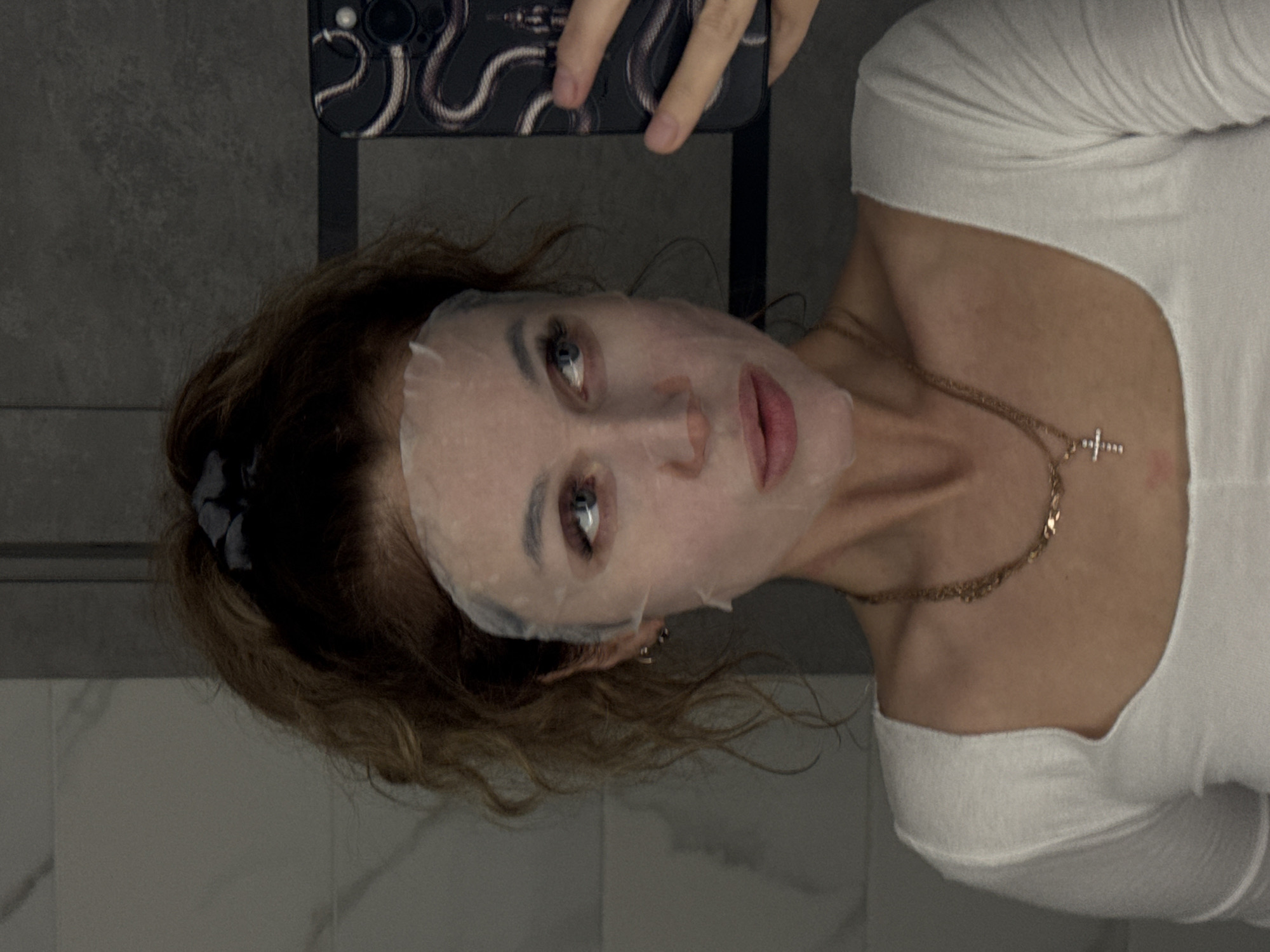 The image shows a woman with a sheet mask on her face, taking a selfie in a bathroom. She has wavy, reddish-brown hair pulled back in a ponytail and is wearing a white top and a gold necklace with a cross pendant. Her eyes are blue and she is looking to the side. Her phone, which has a snake-patterned case, is visible in her hand. The background appears to be a tiled wall, suggesting a bathroom setting. The lighting is dim, with some shadows cast on the wall. There is no discernible information within the image to associate it with Chaikovsky, Russia, or any specific city.