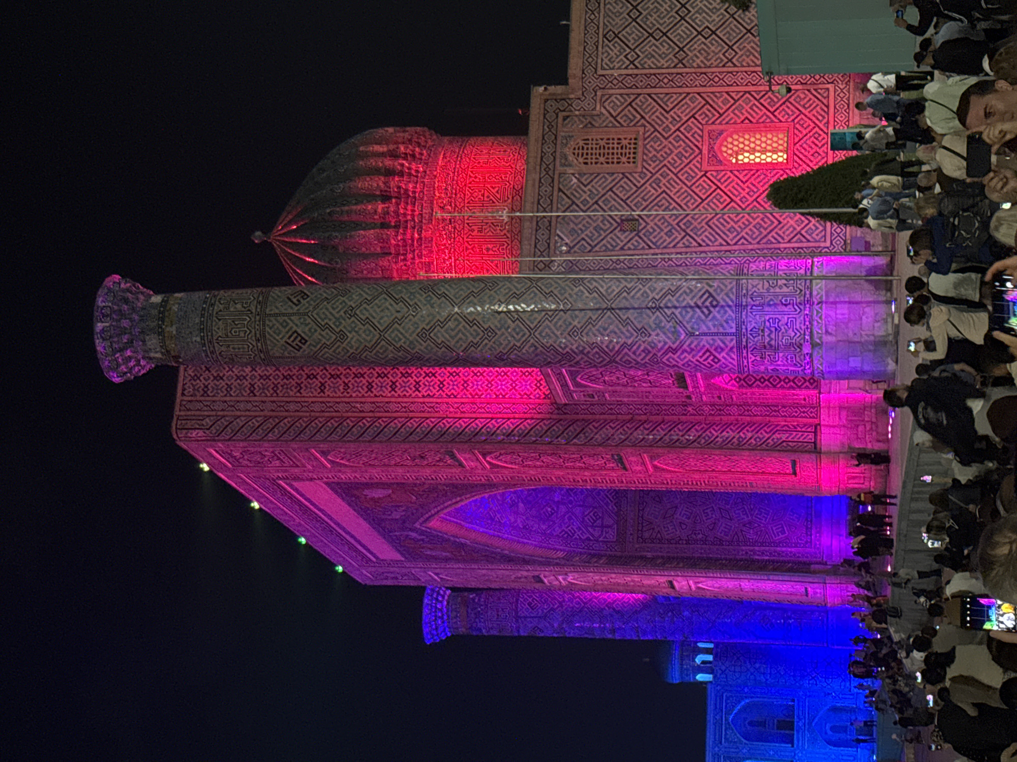 At night in Samarkand City, Uzbekistan, a large architectural structure, likely a historical building or monument, is illuminated with colorful lights. The top of the structure is lit in red, transitioning down to shades of purple and blue. The building features intricate patterns and designs. A crowd of people is gathered on the right side of the frame, many of whom are holding up phones or cameras, presumably to take pictures of the illuminated structure. The background is mostly dark, highlighting the building and the crowd.