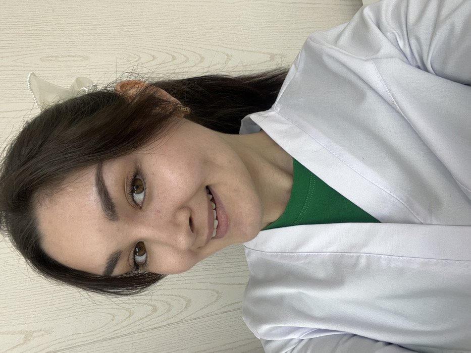A young woman with brown hair, fair skin, and brown eyes smiles. She wears a white lab coat over a green shirt. A white bow accessory is visible in her hair. The background appears to be a light-colored, wood-grain wall. The photo is slightly rotated.
