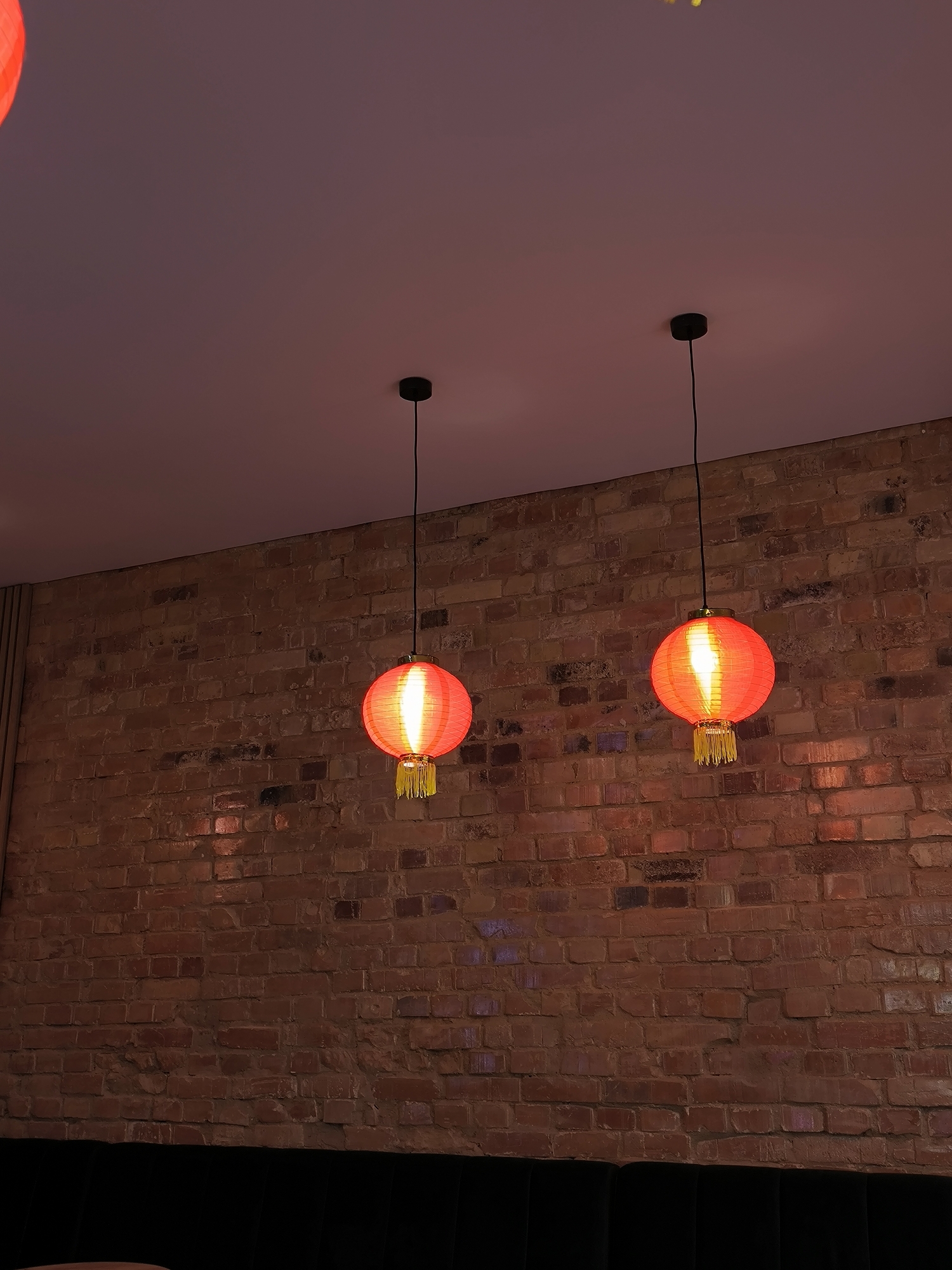 The image shows two red Chinese lanterns hanging from a ceiling in front of a brick wall. The ceiling is pale pink. The lanterns are illuminated and have yellow tassels. The brick wall is a mix of red and brown bricks with visible mortar. A dark object, possibly a piece of furniture, is partially visible at the bottom of the image. There is no indication in the image that identifies the location as Vinnytsia, Ukraine.