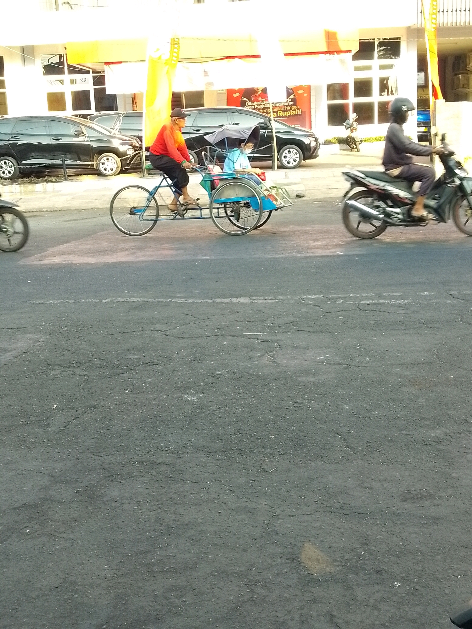 This is a medium-shot, eye-level, outdoor, full-color photograph of a street scene in Jember, Indonesia. In the foreground, the dark asphalt of a road with faint white lane markings is visible. The middle ground shows a pedicab with a man in an orange shirt pedaling. Behind him, a child in a light blue shirt sits in the pedicab. To the right of the pedicab, a person wearing a dark jacket and helmet rides a black and silver motorcycle. In the background, several black cars are parked on the side of the road, and a white building with large windows and yellow awnings stands behind them. The overall lighting suggests it is daytime, possibly late afternoon.
