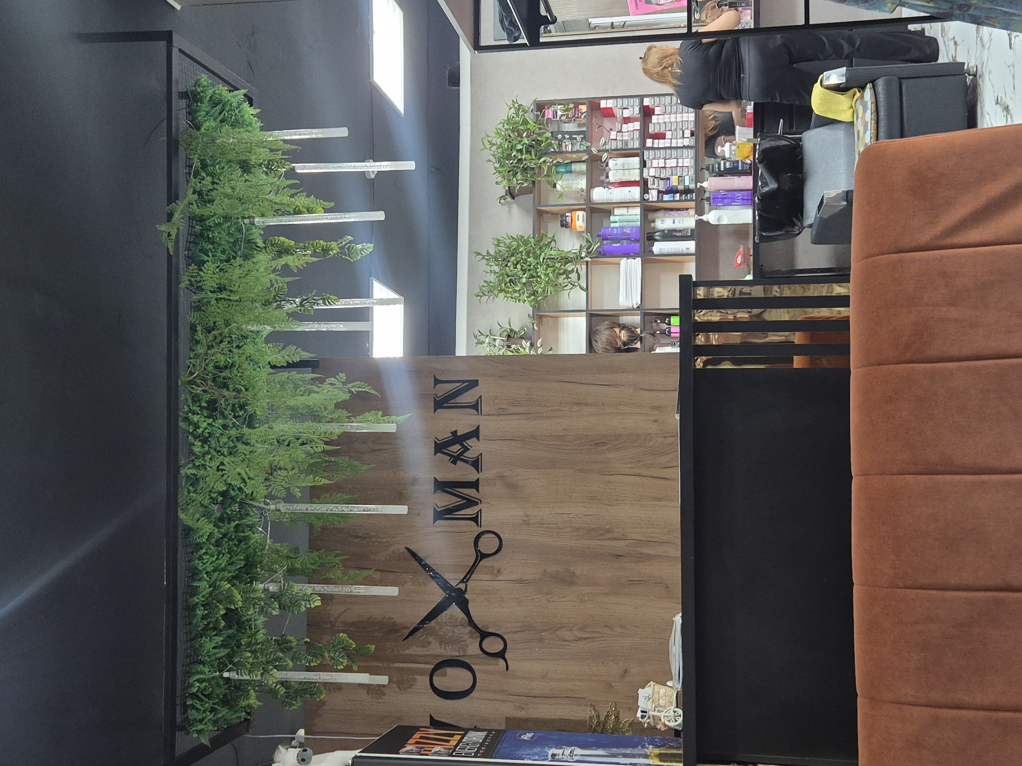 The image depicts the interior of a salon, likely a hair salon, located in Kokshetau, Kazakhstan. The foreground features a section of a plush, rust-colored sofa. In the midground, a black reception desk is visible, behind which a wooden panel is mounted on the wall. This panel is adorned with a large, black silhouette of scissors overlaid with the stylized text "Y O MAN." Hanging from the ceiling above the reception area are artificial green plants and clear cylindrical light fixtures. To the right of the reception desk, shelves are stocked with various hair care products, bottles, and boxes, indicating a retail or service area. A hair washing station with a black sink and chair is also present. In the background, a woman with blonde hair, wearing black attire, is standing and appears to be engaged in some activity at the shelves. The overall environment suggests a modern and clean salon setting.