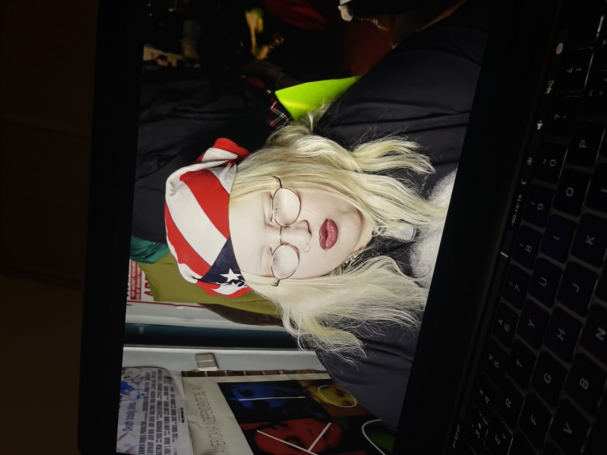 The image displays a screen showing a person, with a laptop keyboard partially visible in the foreground to the right. The screen is oriented sideways.
The person on the screen has pale skin, long, light blonde hair, and is wearing round, wire-frame glasses and red lipstick. A bandana with a prominent red and white stripe pattern, resembling a section of the US flag, covers part of their head. They are dressed in dark clothing, with a flash of bright neon green or yellow fabric visible around their neck area. The person's eyes are partially closed or cast downwards, giving them a pensive or serene expression.
The background behind the person on the screen is dark and indistinct. Below the displayed person, on what appears to be a wall or surface within the displayed content, are posters or papers. Partially visible text on these items includes "Deat" and "AMERICANA NDEPENDENTE DE".
To the right, the black keys of a laptop keyboard are visible, along with parts of the laptop's bezel. Timecodes "01:53:01" and "-00:14:16" are visible on the screen's edge, suggesting the display shows a video. The overall lighting of the physical environment where the photograph was taken is dim, with the screen providing the main illumination. The location context for the image is Torrent, Spain, although no specific visual cues in the image confirm this.