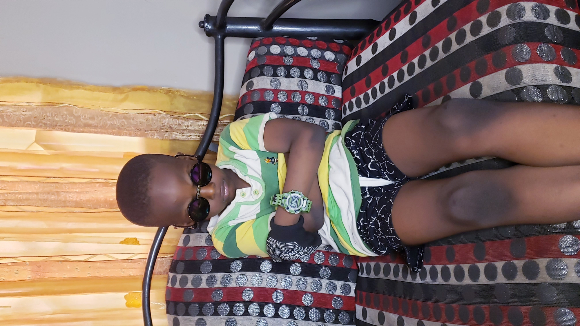 A young person lies on a padded chair in what appears to be a home setting. The child, wearing dark sunglasses, a striped shirt, patterned shorts, a watch, and a glove on one hand, is reclining on a seat with striped and polka-dot cushions. The background includes yellow curtains and a metal frame from the furniture. This scene occurs in Narayi, Nigeria.